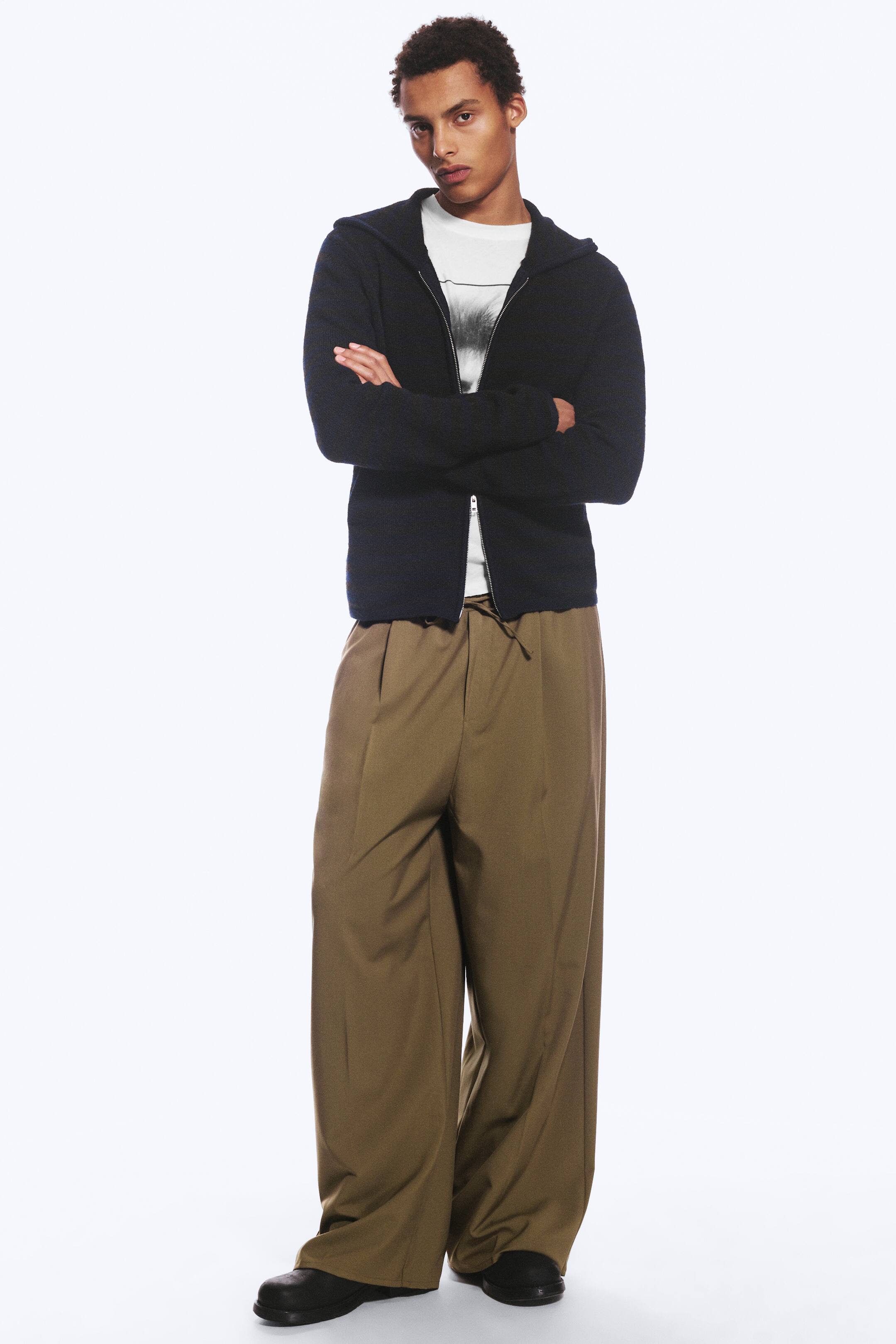 Преглед на по-голямо изображение: Моделът е облечен с широки, oversized костюмни панталони в каки-кафяво, с връзка на талията и изгладени ръбове. Панталоните са стилизирани с бяла тениска с графичен принт, тъмно суичър яке с цип и качулка и черни обувки.