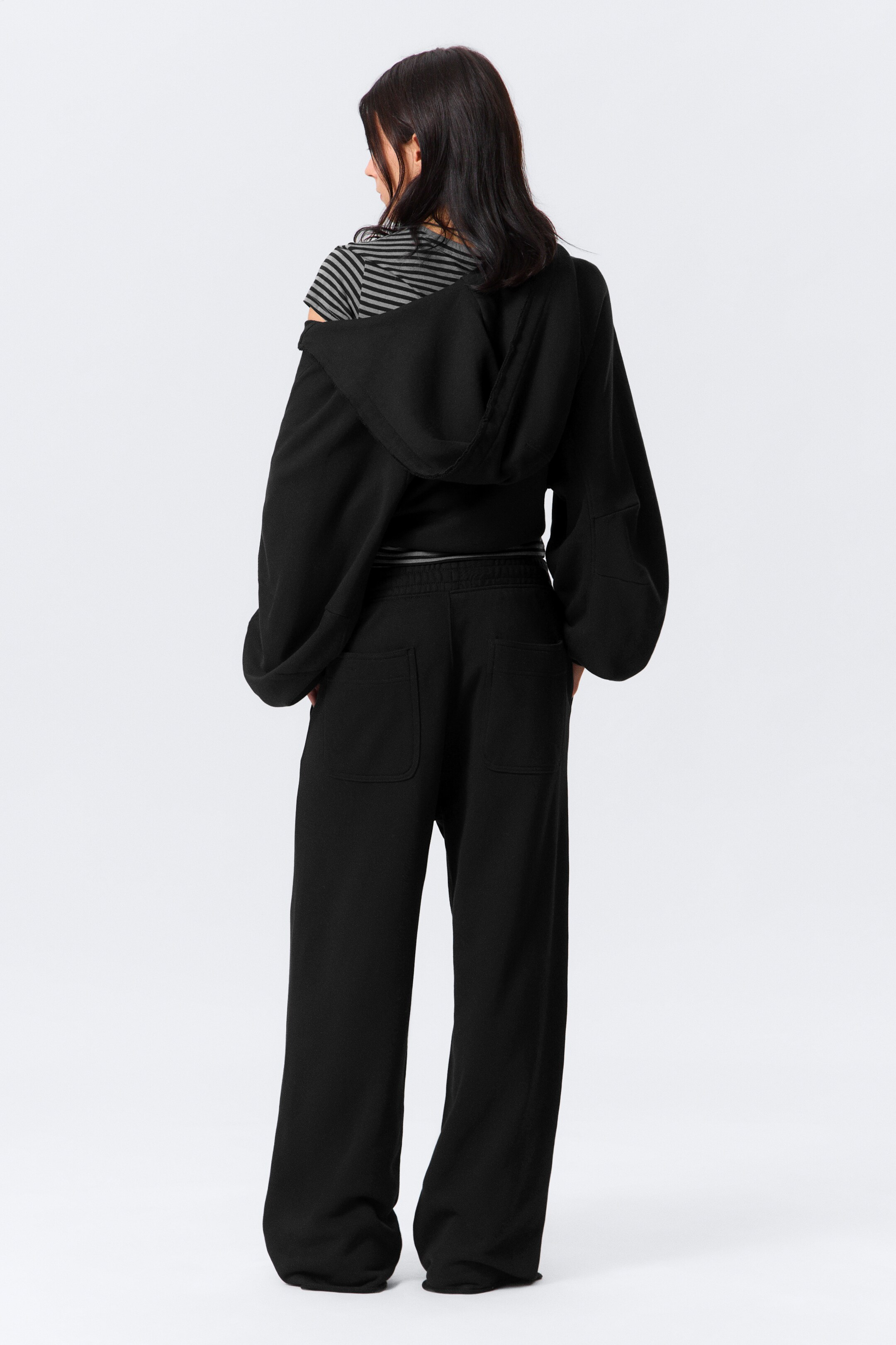 Agrandir l'image: Un ensemble de détente noir est présenté de dos, comprenant un haut à capuche avec des manches volumineuses et un pantalon à jambes larges. Le haut est porté sur une épaule, révélant un t-shirt rayé en dessous. Le pantalon est conçu avec des poches arrière plaquées et une taille élastiquée.
