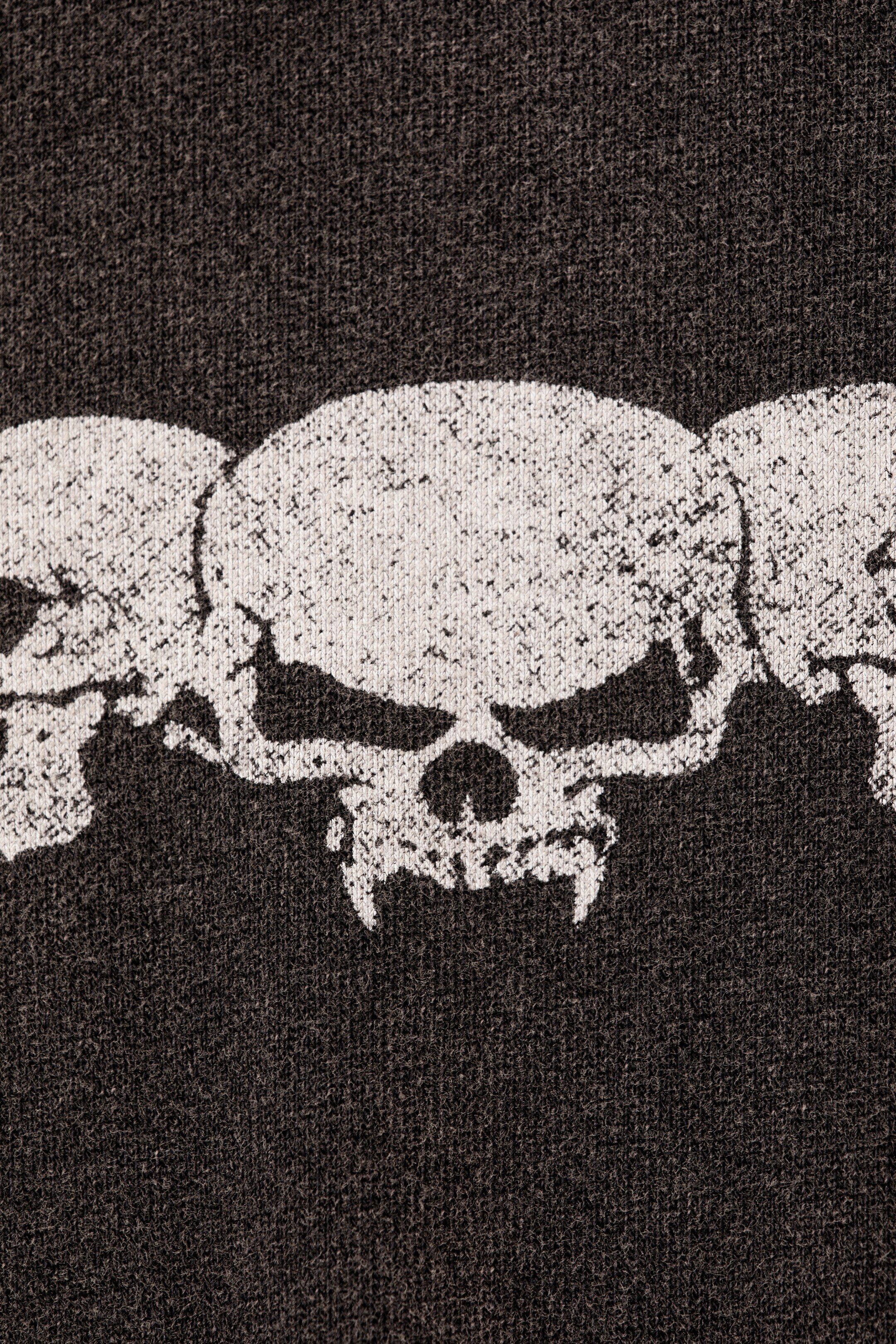 Ingrandisci l'immagine: Una T-shirt a maniche lunghe e squadrata mostra un primo piano del suo davanti, mettendo in risalto una stampa grafica di tre teschi bianchi stilizzati con un effetto invecchiato su un tessuto scuro e strutturato. Il teschio centrale presenta zanne prominenti, con altri due teschi parzialmente visibili su entrambi i lati, disposti orizzontalmente.