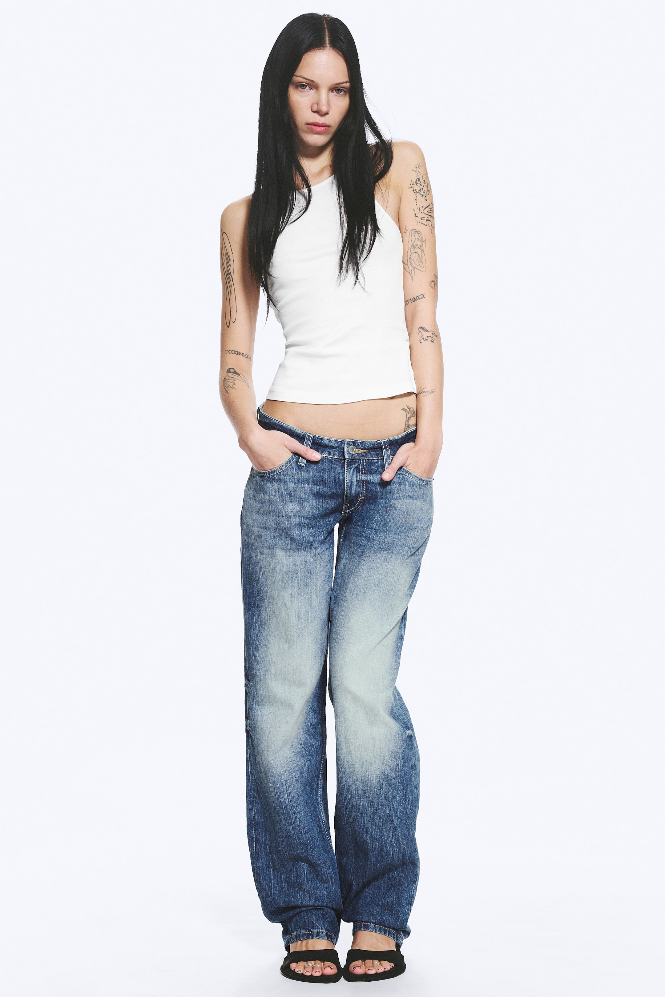 Grotere afbeelding bekijken: Een witte geribbelde tanktop met een hoge halslijn en een aansluitende pasvorm. Het wordt gedragen met laaghangende blauwe wijde jeans, die een vervaagde wassing tonen met lichtere dijen en een wijde pijp, die bij de enkels samenkomt.