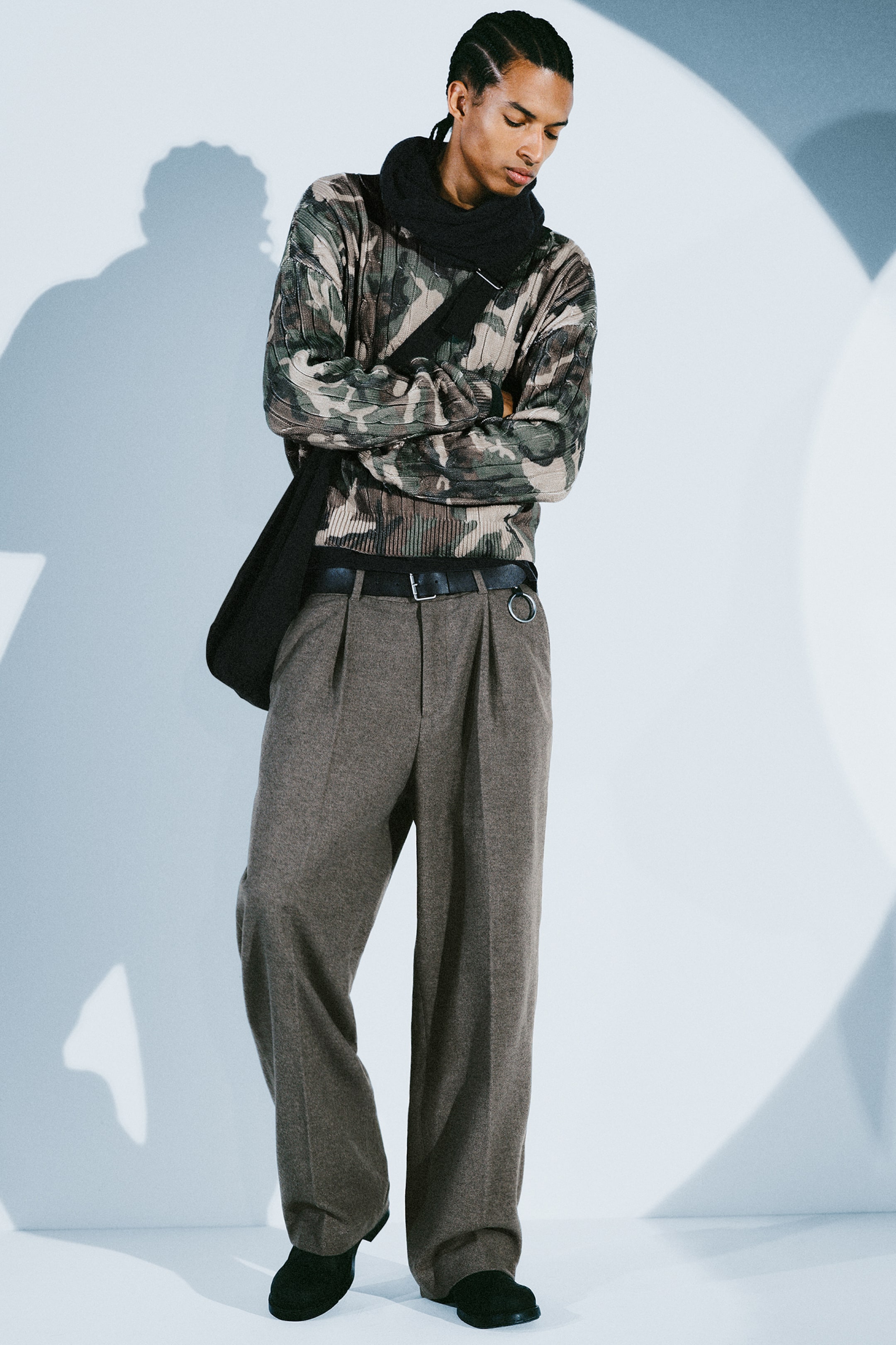 View larger image: A pair of brown wool-blend tailored trousers featuring front pleats, pressed creases, and a relaxed wide-leg silhouette is worn by the model. The trousers are styled with a black leather belt, a camouflage-patterned jumper, and black shoes.