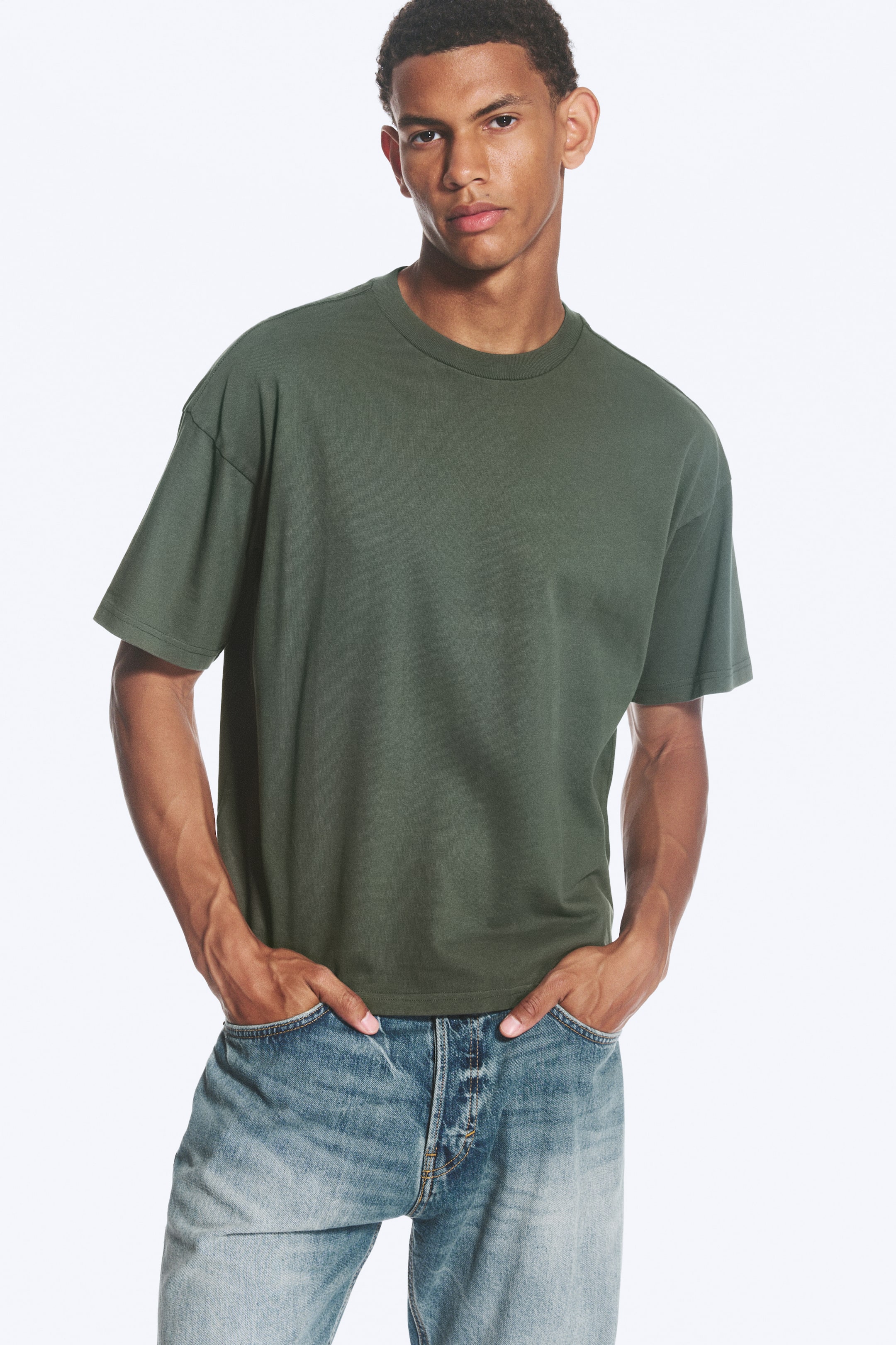 Agrandir l'image: Un T-shirt oversize vert foncé est porté par le mannequin, avec un col rond, des épaules tombantes et des manches larges. Le T-shirt présente une coupe ample alors que le mannequin pose les mains dans les poches.