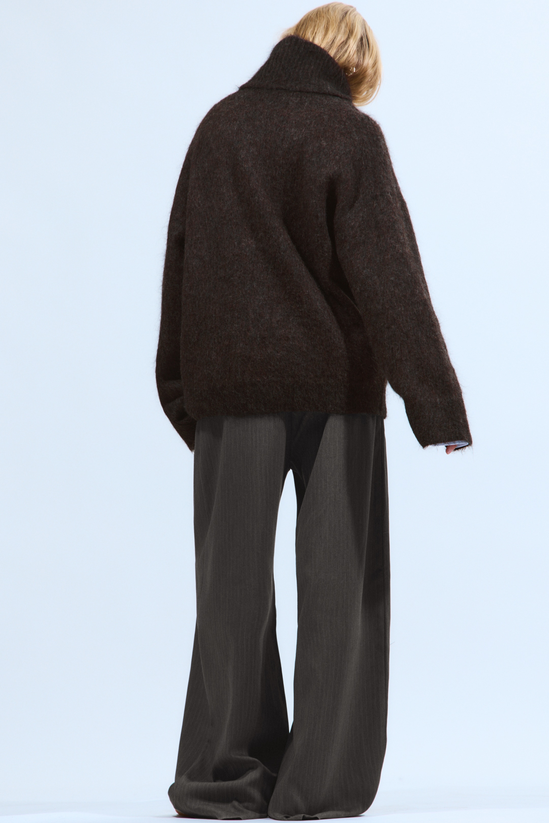 Größeres Bild anzeigen: Ein dunkelbrauner, flauschiger Strickrollkragenpullover mit übergroßer Passform und langen, weiten Ärmeln wird von hinten gezeigt, wobei ein hellblaues Bündchen unter einem Ärmel hervorschaut. Er wird mit dunkelgrauen, weit geschnittenen Hosen kombiniert, die subtile vertikale Streifen aufweisen und auf dem Boden aufliegen.