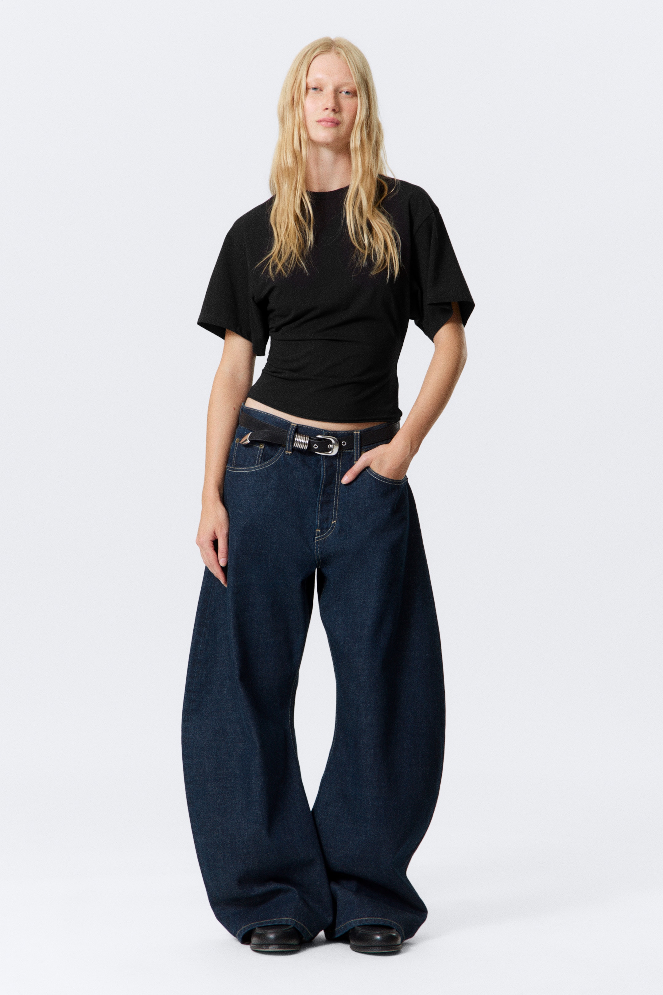 Grösseres Bild ansehen: Ein schwarzes, kurzärmeliges Oberteil wird präsentiert, das einen Rundhalsausschnitt und eine taillierte Passform aufweist. Dunkel gewaschene, weit geschnittene Jeans mit Kontrastnähten werden gezeigt, kombiniert mit einem schwarzen Gürtel, der eine silberfarbene Schnalle hat.