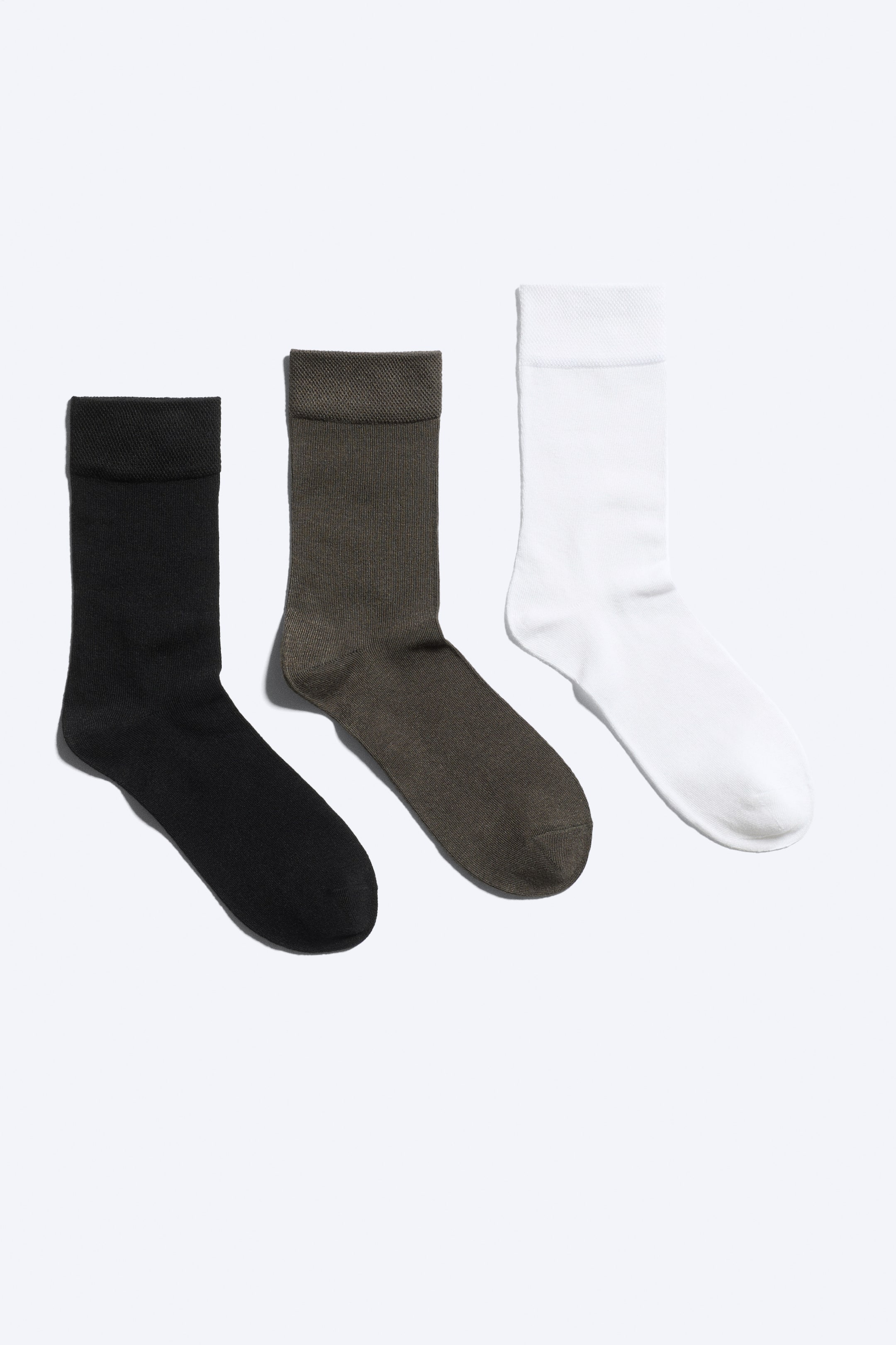 Visa större bild: Ett 3-pack strumpor visar tre enskilda strumpor i svart, mörkbrunt och vitt, placerade sida vid sida. Varje strumpa har en slät, enfärgad stickad kropp och en texturerad, pikéstickad mudd.
