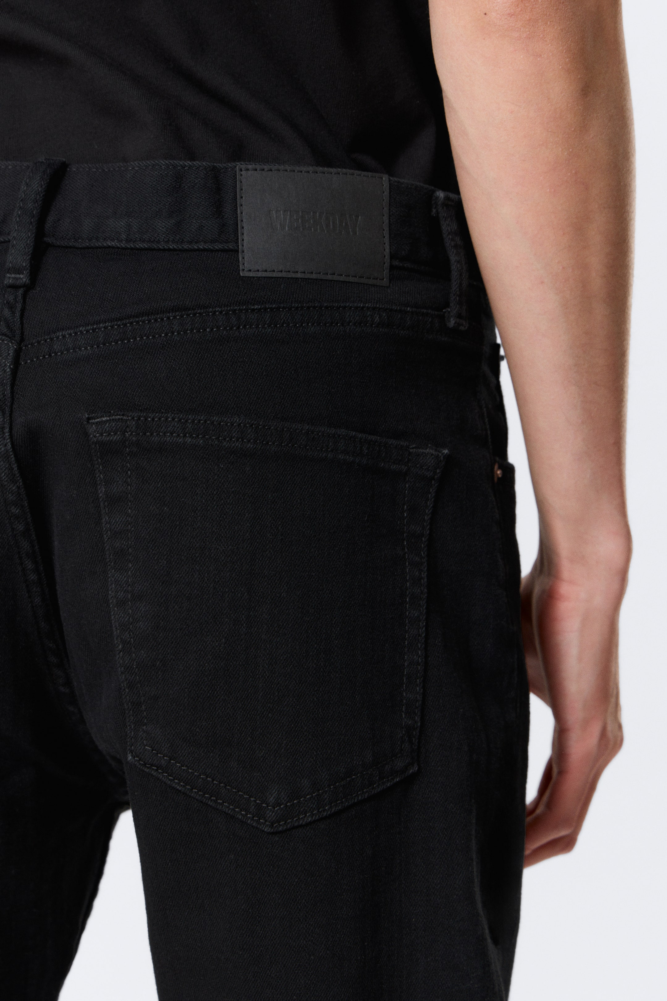 Se større billede: Sorte jeans præsenteres fra en bagfra og let vinklet vinkel, med et rektangulært sort mærke patch med "WEEKDAY" præget på den bageste linning. To store baglommer med tone-i-tone syninger er synlige.
