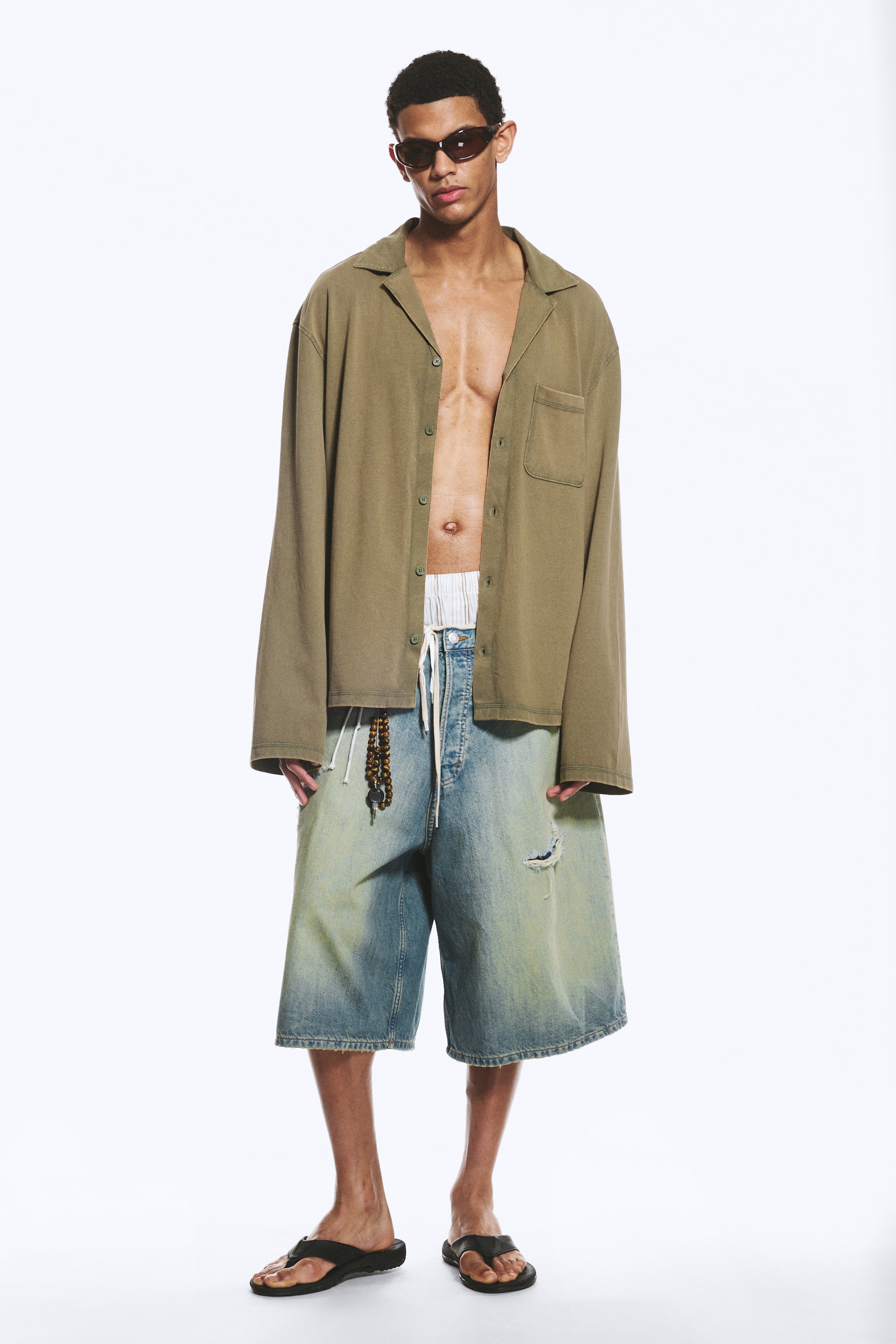 Se større billede: En oversized olivengrøn langærmet skjorte med en camp-krave, knaplukning foran og en enkelt brystlomme bæres åbent af modellen, hvilket afslører et stribet taljebånd.