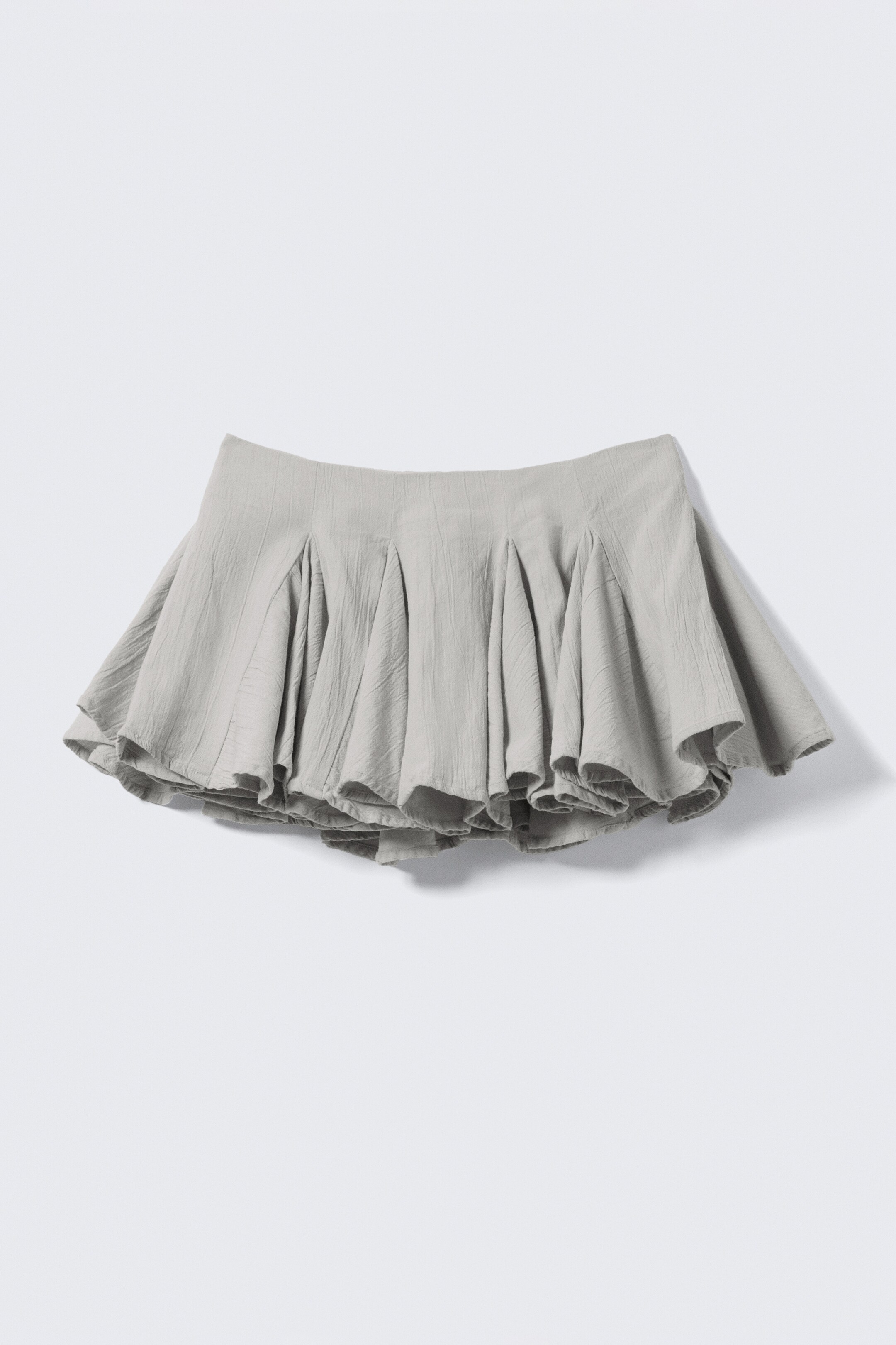 Agrandir l'image: Une jupe mini gris clair est présentée de face, avec une ceinture lisse. Elle affiche une silhouette volumineuse et plissée avec une forme évasée et une structure subtile dans son tissu texturé.