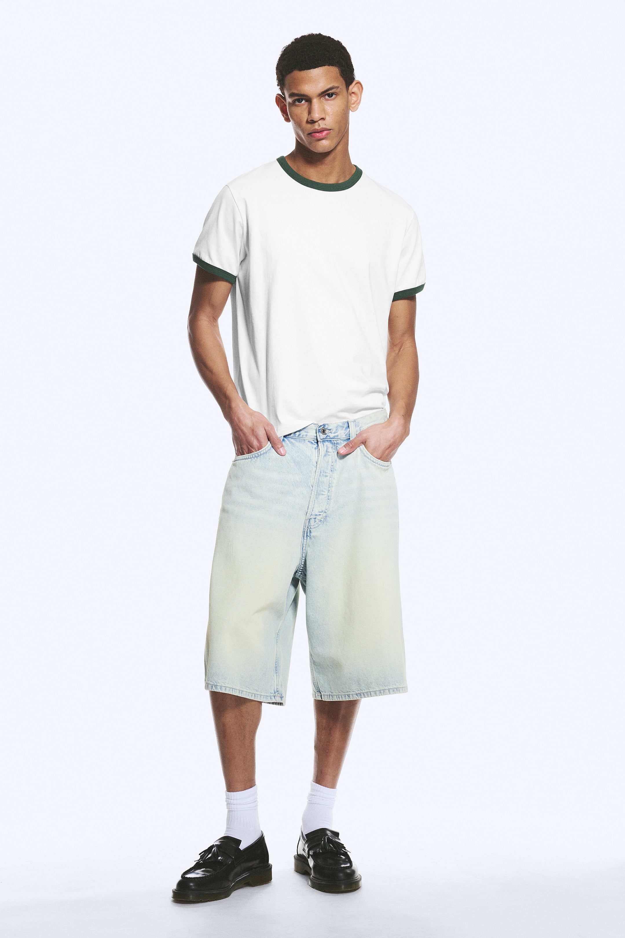 Vis større bilde: Modellen har på seg mid-rise, lysvaskede denimshorts med en løs, baggy passform, og har hendene i frontlommene. Shortsene har et klassisk femlommers design og oppbrettede kanter.