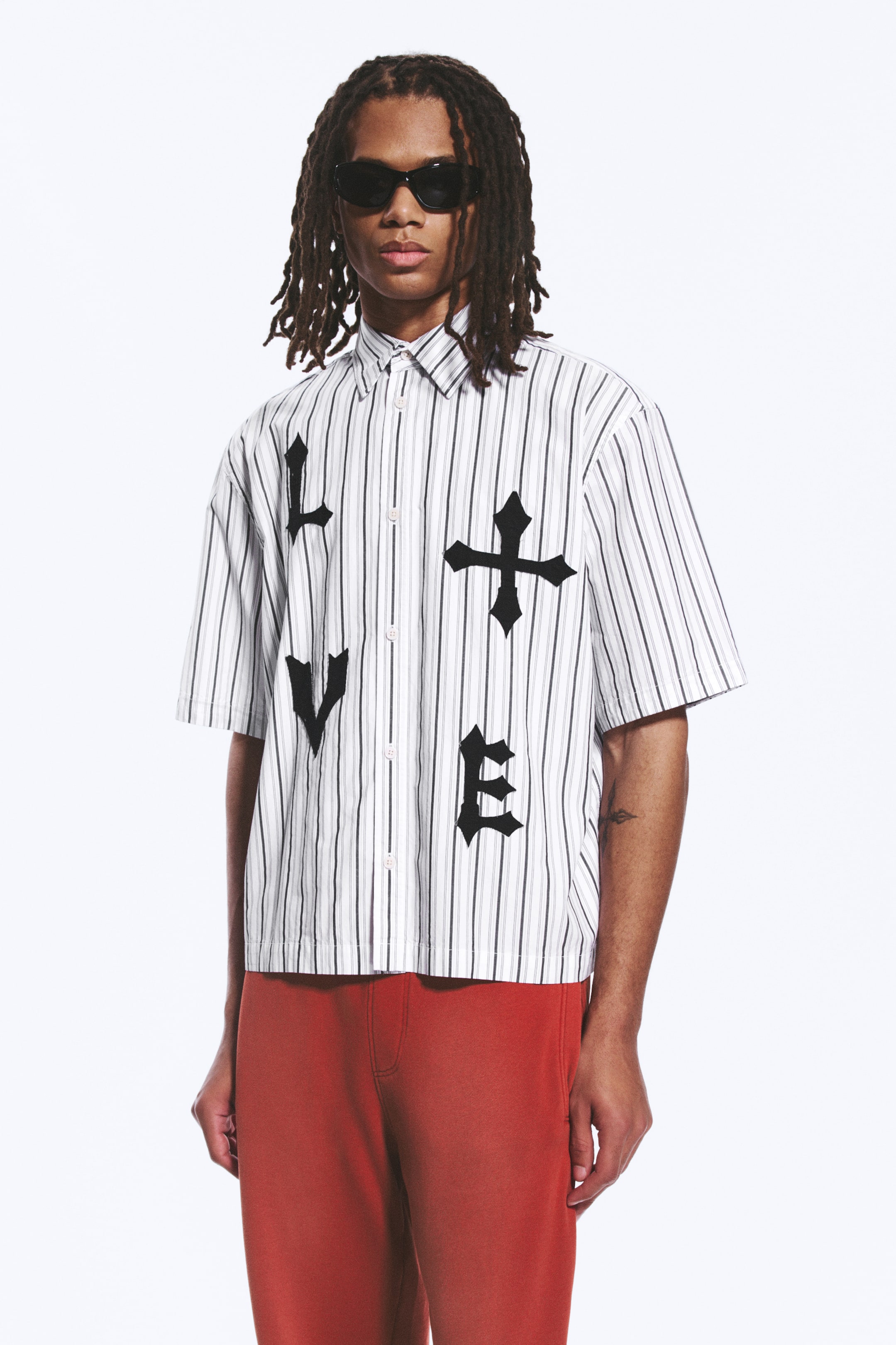 Agrandir l'image: Une chemise à manches courtes à boutons présente des rayures verticales noires et grises sur un fond blanc. De grandes lettres gothiques noires forment "L", "V" et "E", avec un symbole de croix gothique complétant le mot "LOVE" sur le devant.