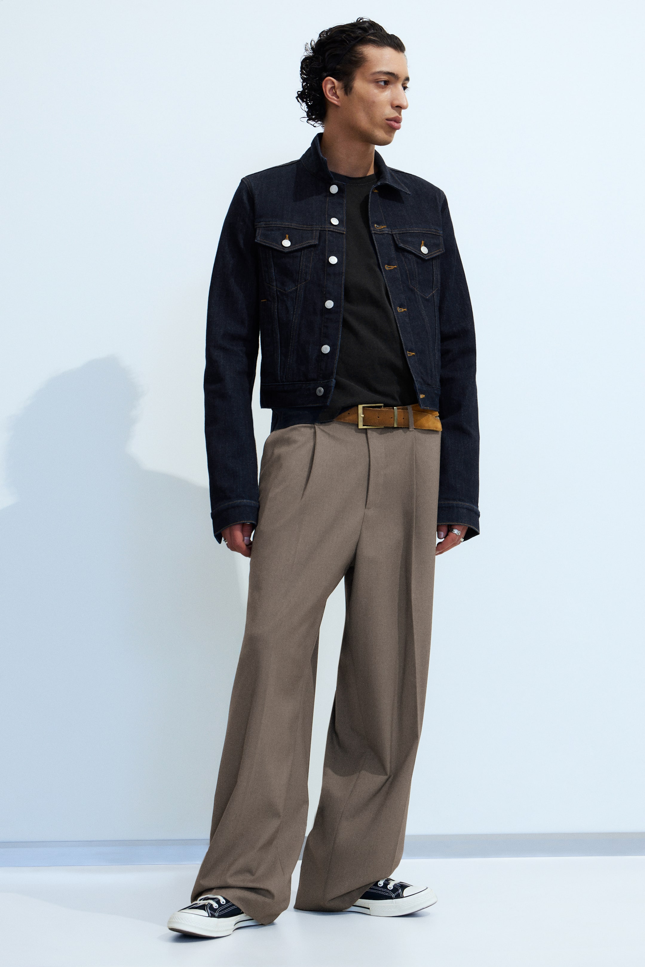 Agrandir l'image: Une veste en jean à délavage foncé avec des boutons argentés et des poches poitrine est portée ouverte sur un t-shirt noir à col rond. Un pantalon large plissé marron est porté avec une ceinture en daim marron avec une boucle dorée, recouvrant des baskets montantes en toile noire.