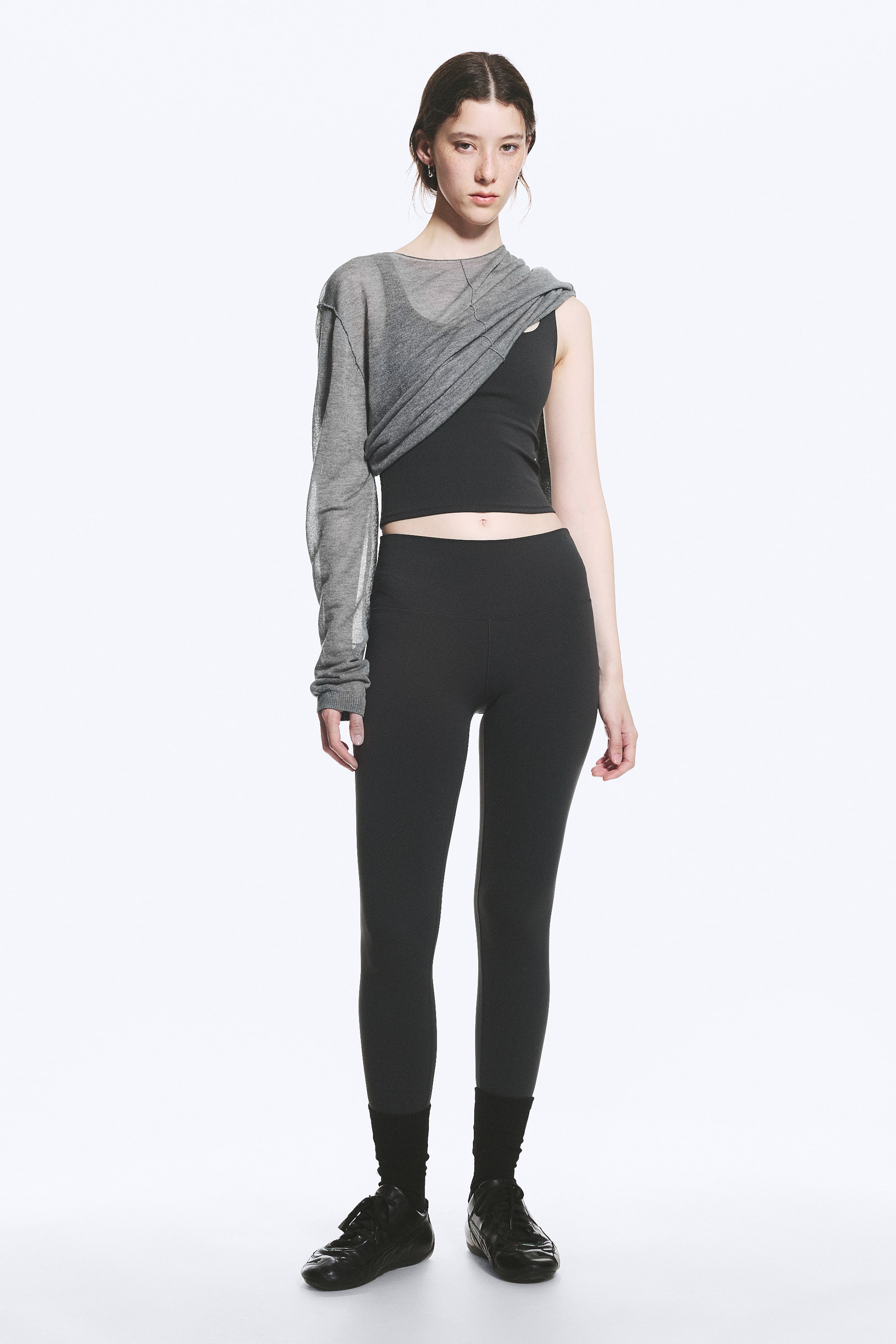 View larger image: A grey sheer knit top featuring a single long sleeve and a wide neckline, draping diagonally across the torso. It is layered over a black cropped tank top, which is paired with black high-waisted leggings.