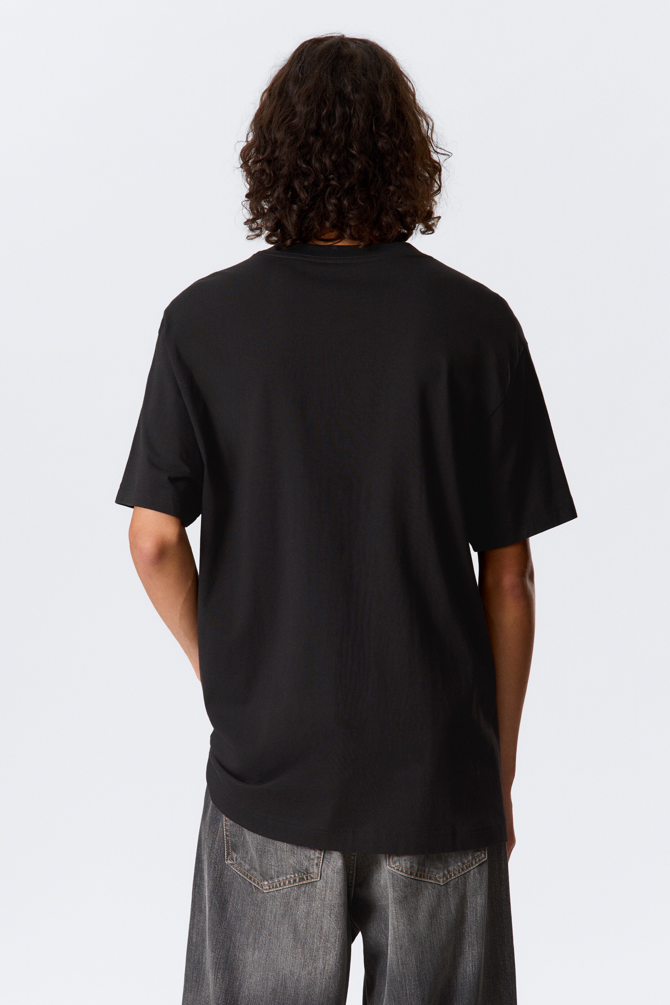 Agrandir l'image: Un T-shirt noir oversize, fabriqué en jersey de coton lourd et doux, est porté par le modèle. Il présente une coupe extra ample, un col rond côtelé, des épaules tombantes et des manches larges.