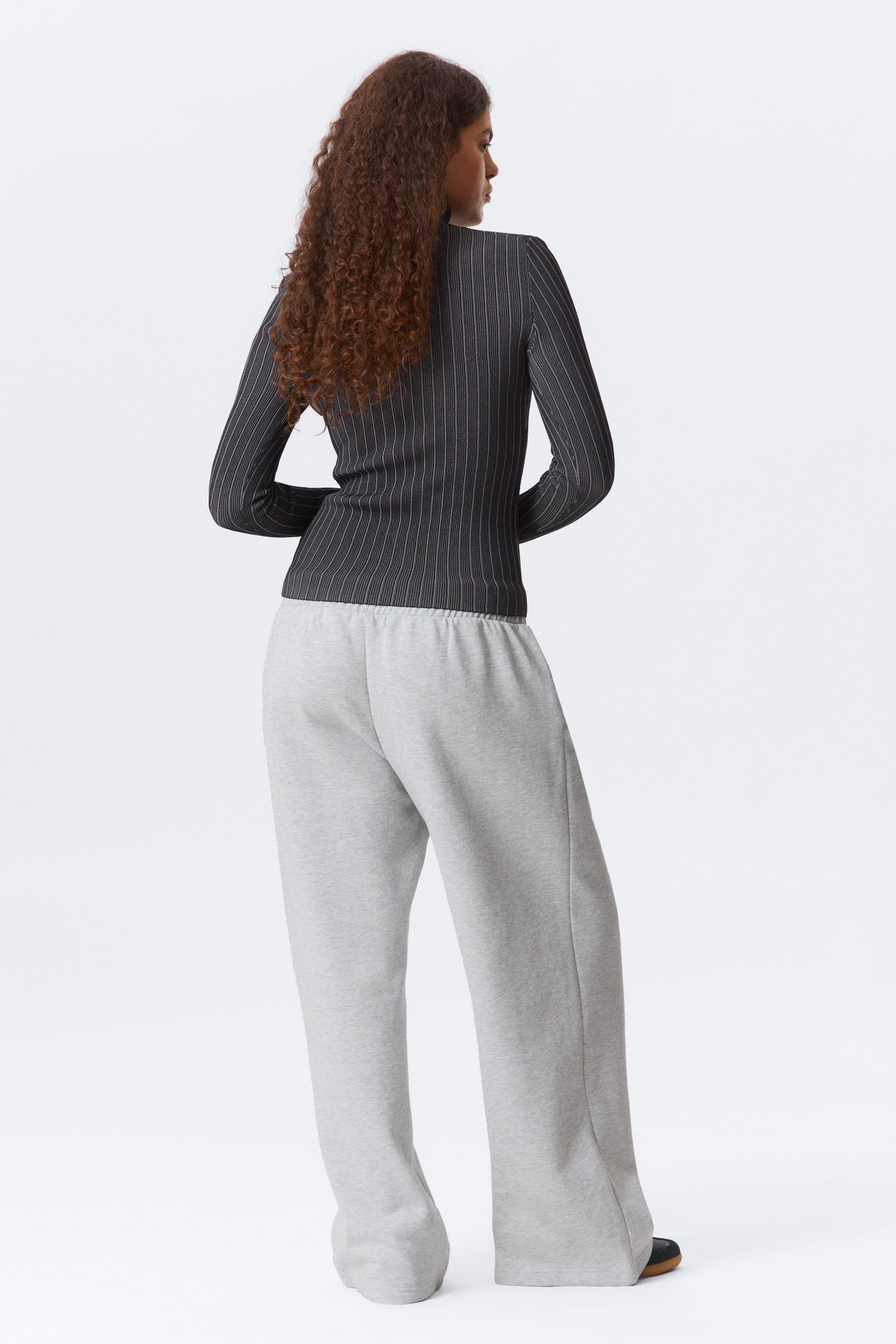 Agrandir l'image: Un haut à manches longues en maille côtelée gris foncé avec des rayures verticales blanches est montré de dos, s'ajustant étroitement au corps. Il est associé à un pantalon de survêtement gris clair à jambes larges, doté d'une taille élastiquée et qui se regroupe aux chevilles.