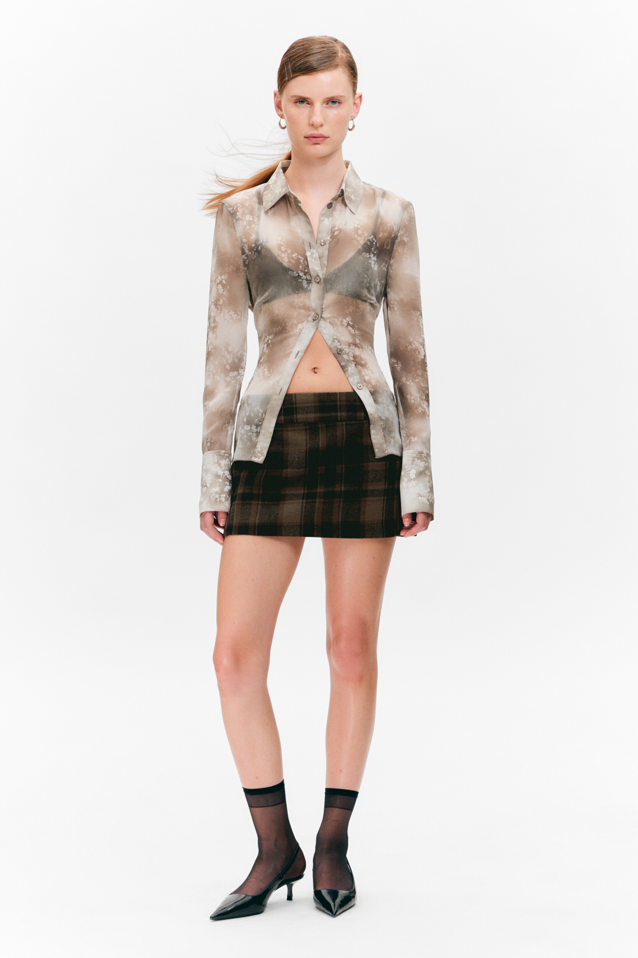 View larger image: A sheer, long-sleeved button-up shirt featuring a subtle floral pattern is being presented from a front angle, showing its fitted silhouette. It is partially unbuttoned, revealing a bra underneath, and is paired with a dark plaid mini skirt. Black sheer ankle socks are being worn with pointed-toe slingback kitten heels.