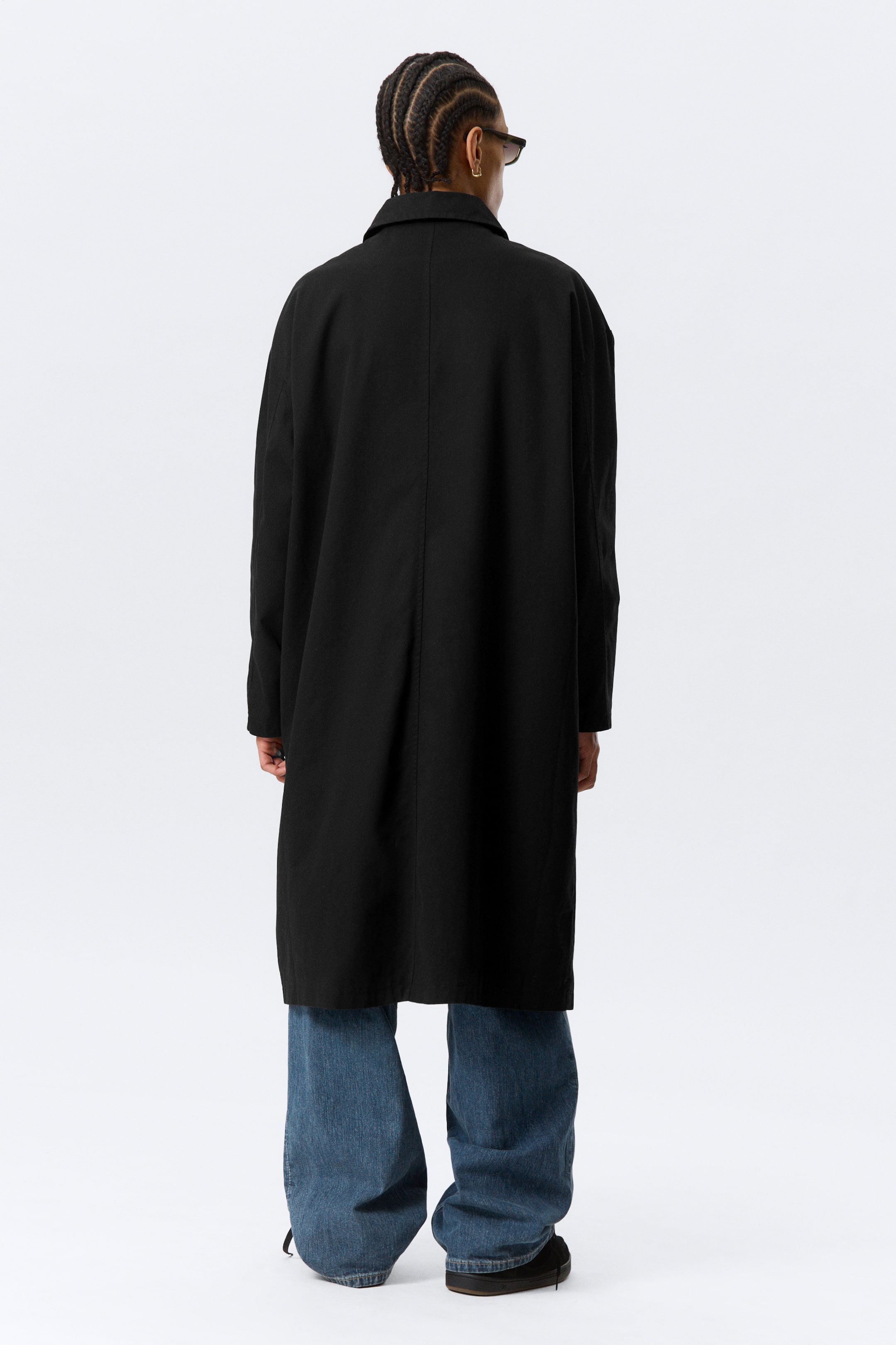 Agrandir l'image: Un manteau noir est présenté de dos, avec une coupe ample et des manches longues, doté d'une couture centrale descendant le long de son dos. Il s'étend au-delà des hanches, couvrant la partie supérieure d'un jean bleu.