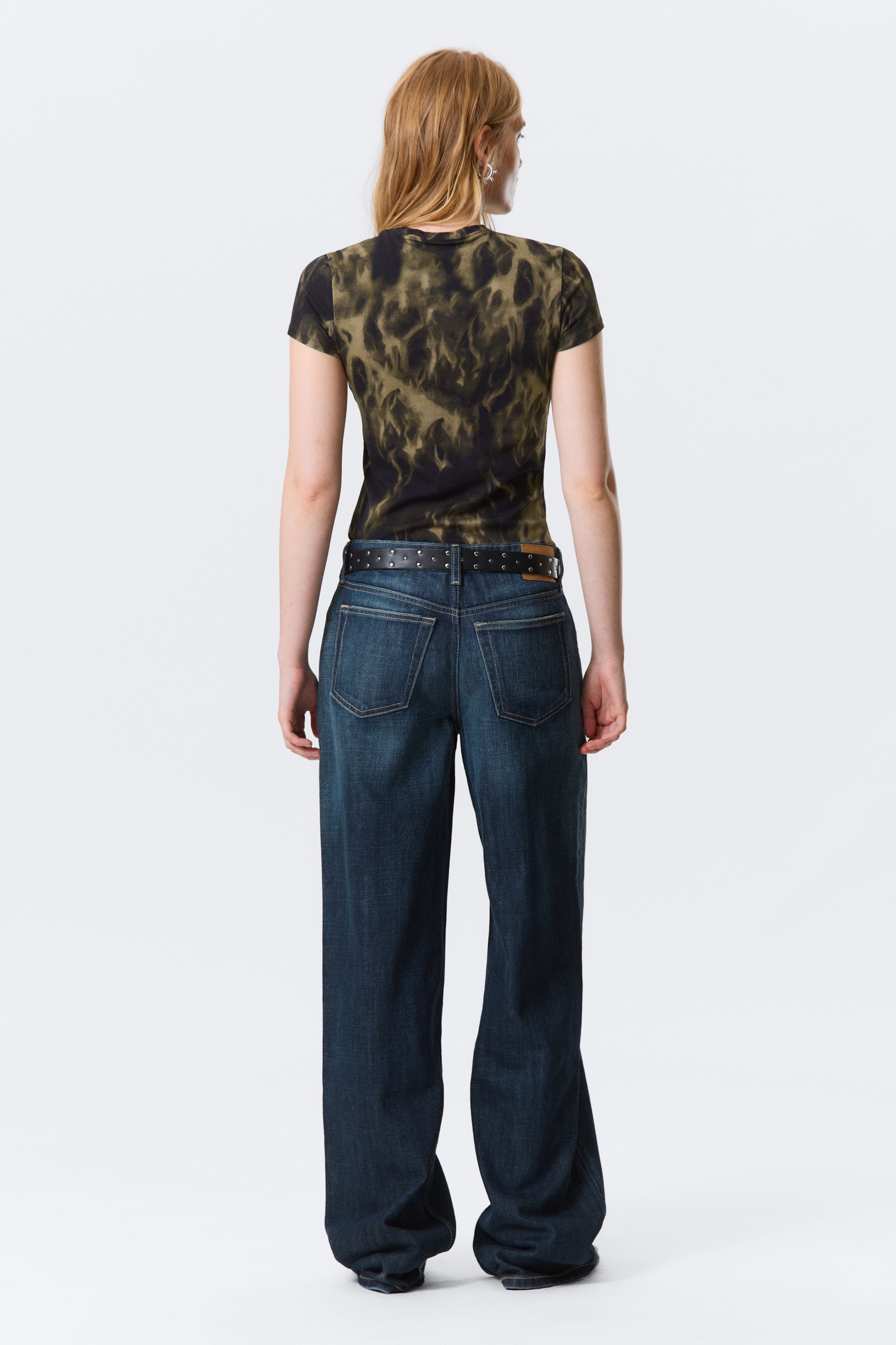 Größeres Bild anzeigen: Ein eng anliegendes, kurzärmeliges T-Shirt wird getragen, das eine dunkle Basis mit einem olivgrünen gesprenkelten Muster aufweist. Dunkel gewaschene, weit geschnittene Jeans werden getragen, gesichert durch einen schwarzen Gürtel mit Nieten, alles von hinten betrachtet.