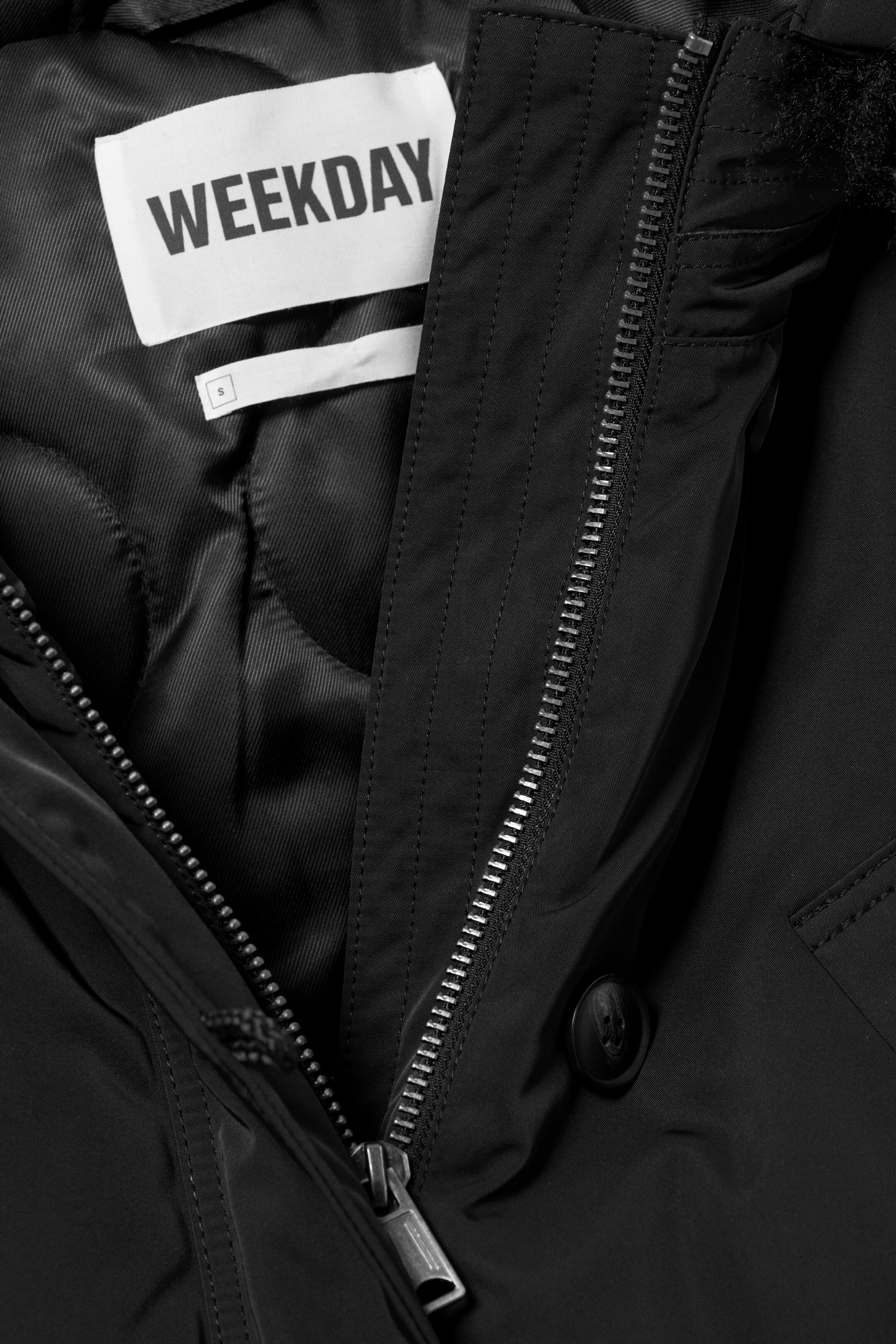 Agrandir l'image: Une veste noire est présentée ouverte, révélant une doublure intérieure matelassée noire. Une étiquette de marque blanche "WEEKDAY" et une plus petite étiquette de taille blanche "S" sont visibles sur la doublure, et une fermeture éclair de ton argenté descend le long de l'ouverture avant de la veste, avec un bouton noir visible à côté.