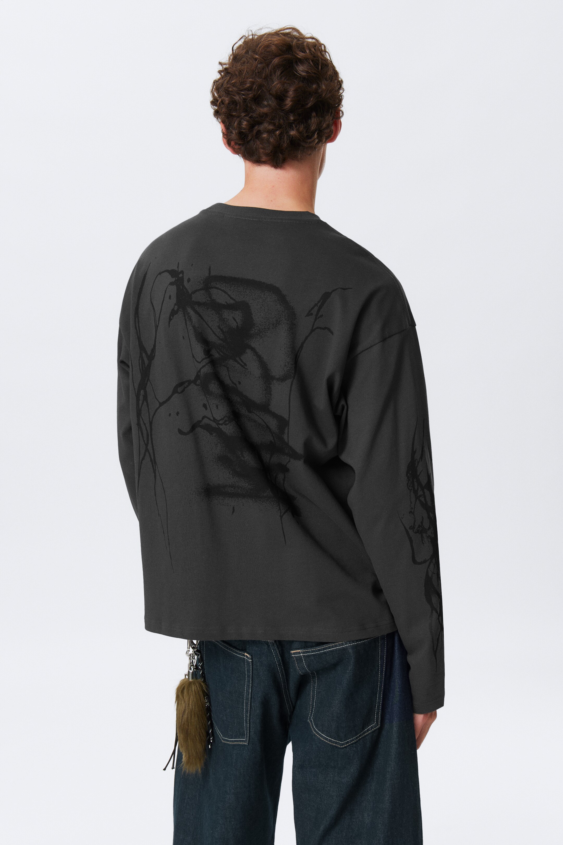 Ingrandisci l'immagine: Una maglia a maniche lunghe grigio scuro oversize è presentata da un'angolazione posteriore, caratterizzata da un grande design grafico astratto nero che copre la parte superiore della schiena e si estende sulle maniche. L'orlo dritto della maglia cade appena sotto la vita, rivelando jeans scuri lavati sotto.