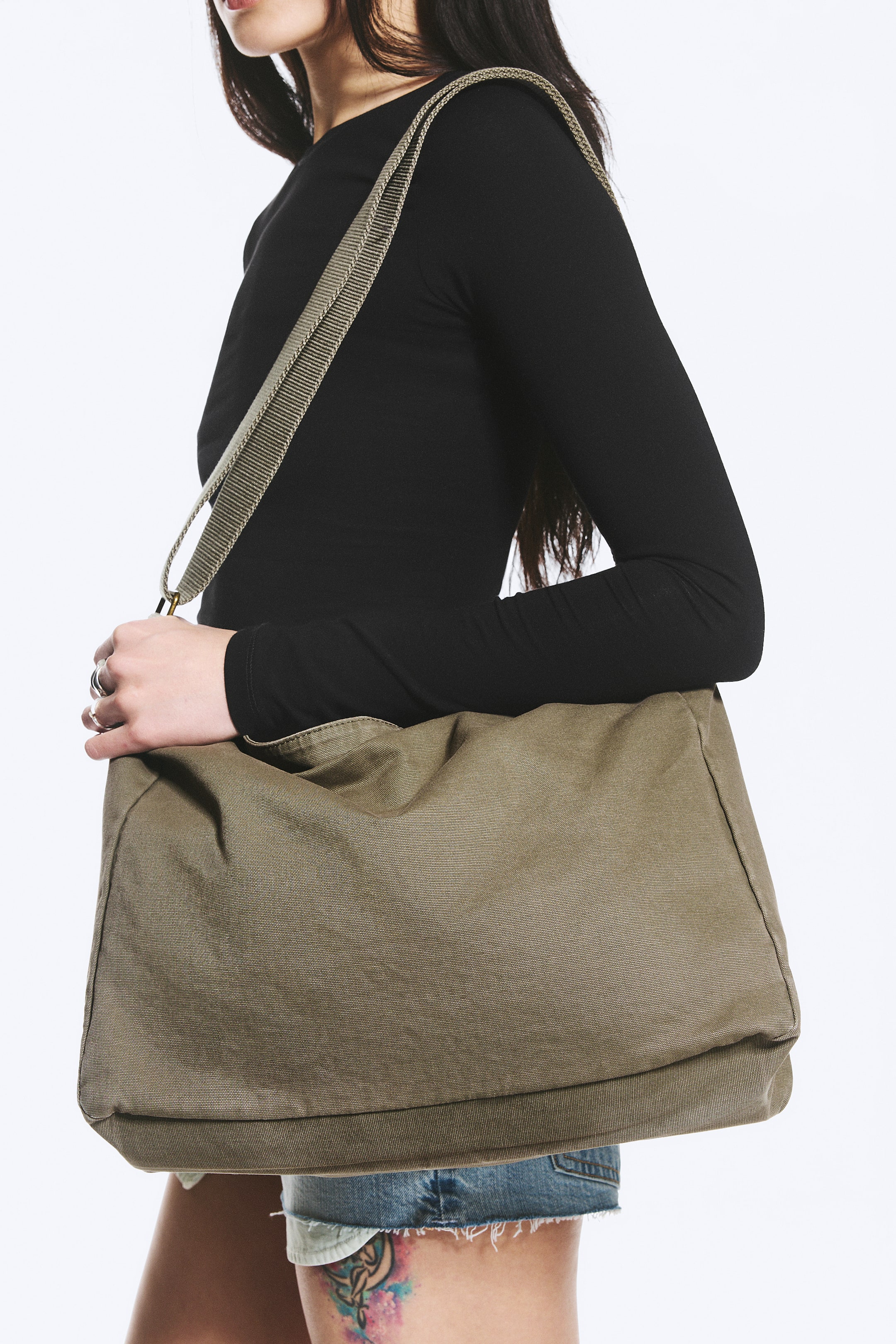 Ver imagem maior: Uma mala de mensageiro em lona verde-azeitona apresenta uma alça larga e texturizada, usada a tiracolo. A mala grande e desestruturada repousa contra a anca, exibindo o seu painel frontal e lateral inferior, com uma mão a segurar o ponto de conexão da alça.