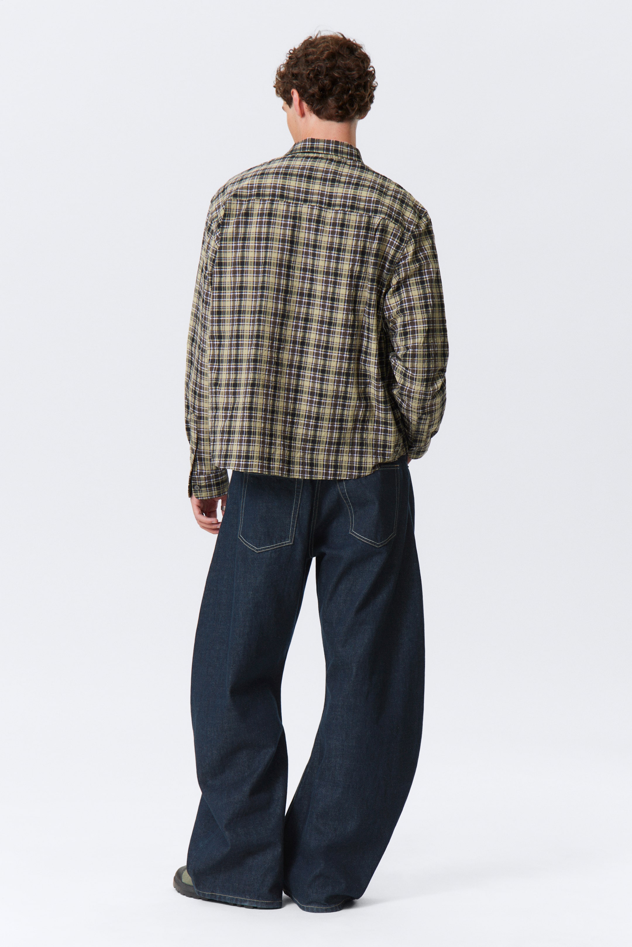 Visa större bild: En rutig skjorta har ett olivgrönt, svart och brunt rutmönster med tunna ljusare linjer, presenterad från en bakre vy med en lös passform. Den bärs med mörktvättade, vida jeans som visar framträdande ljusfärgad sömnad på bakfickorna och sömmarna.