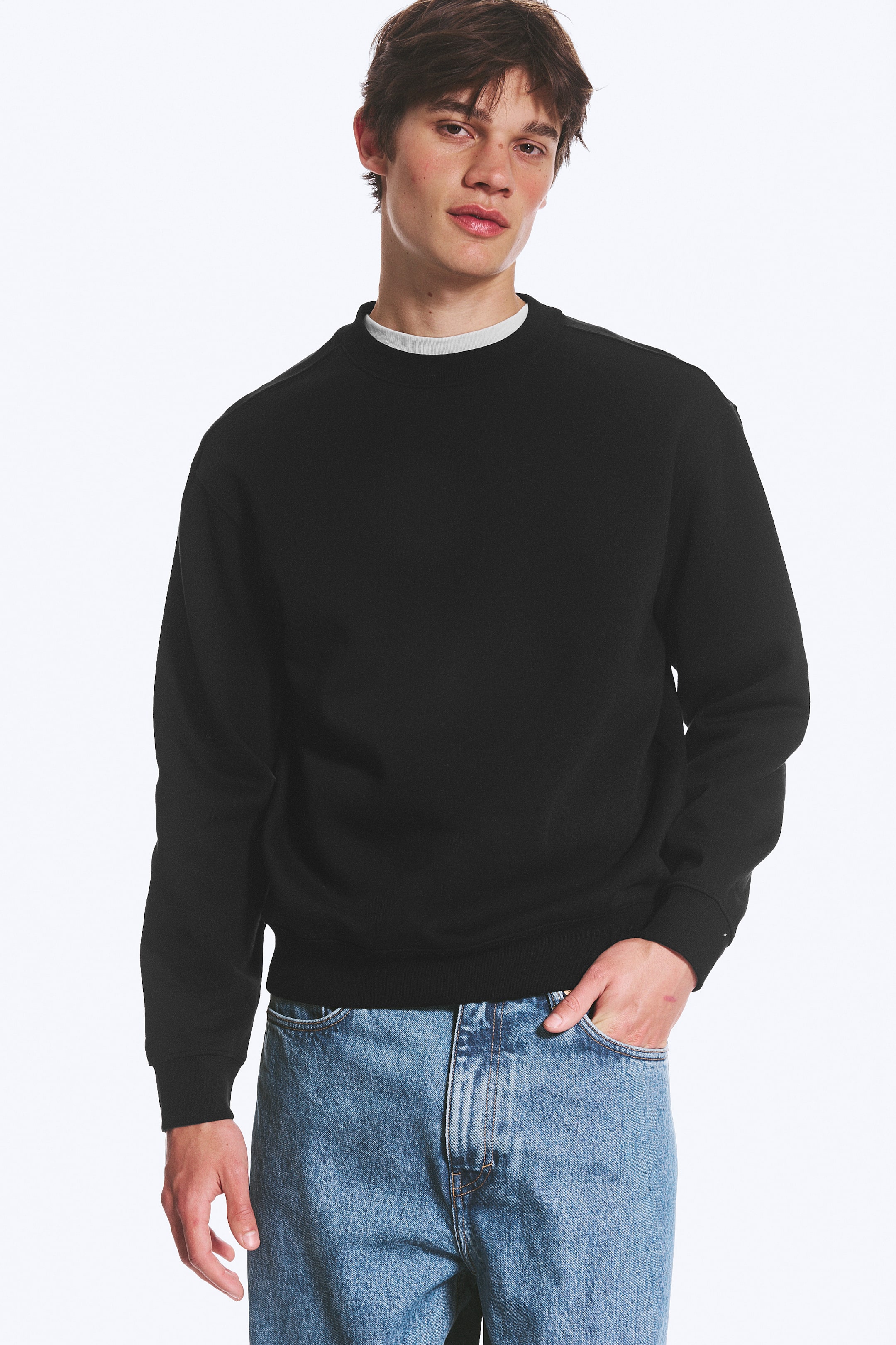 Visualizza immagine più grande: Una felpa nera con scollo tondo presenta una vestibilità comoda, maniche lunghe con polsini a coste e un orlo a coste, leggermente inclinata verso destra. Una t-shirt bianca è visibile sotto la felpa con scollo tondo.
