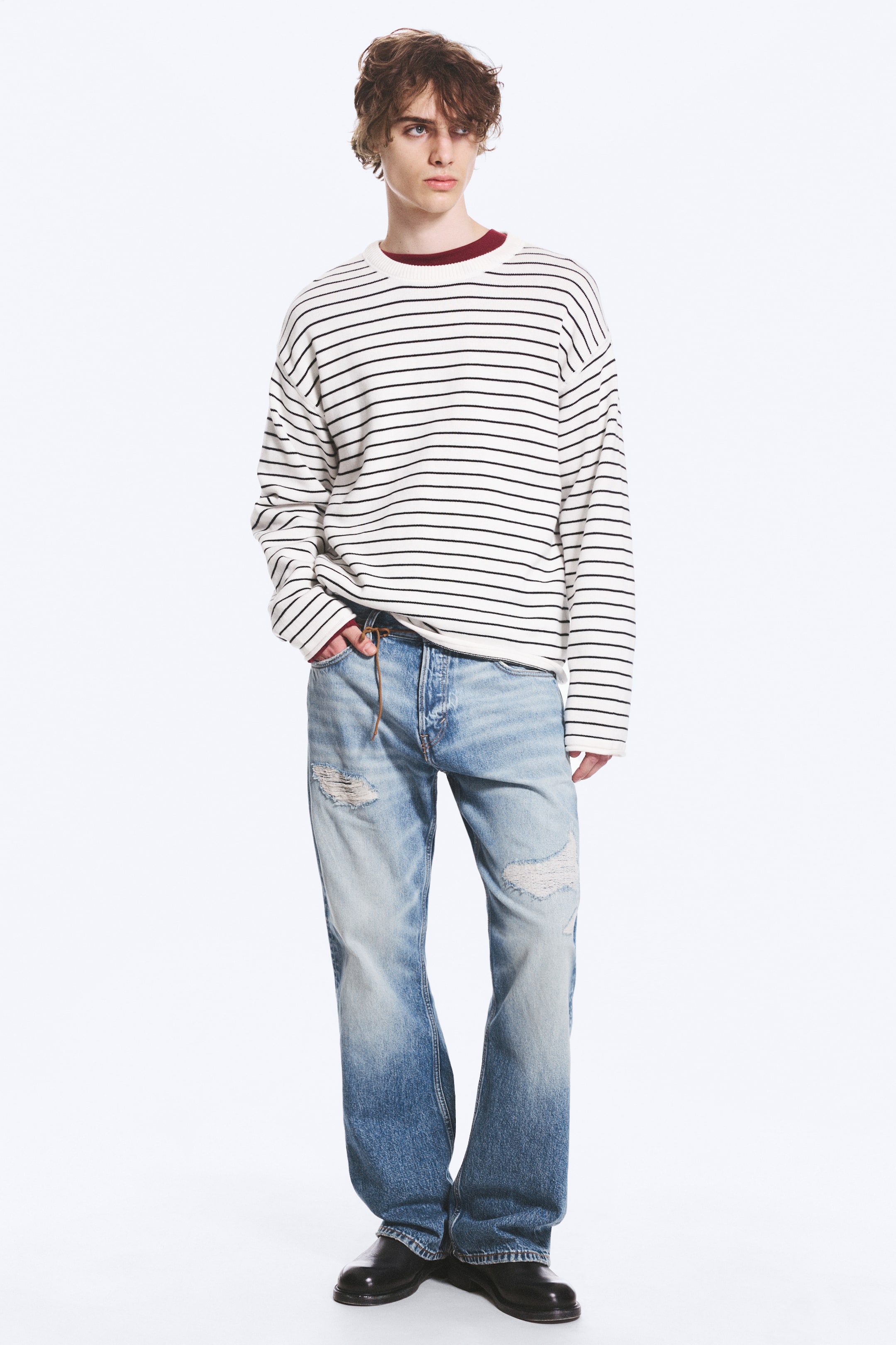 Grösseres Bild ansehen: Ein übergroßer, cremeweißer Pullover mit Rundhalsausschnitt und schwarzen horizontalen Streifen sowie gerippten Bündchen wird über einem dunkelroten Unterhemd getragen und bietet eine entspannte Passform. Dazu kombiniert sind hellblaue, abgenutzte Jeans mit Rissen an den Oberschenkeln und einem dunklen Fade am Saum, zusammen mit schwarzen Lederschuhen.