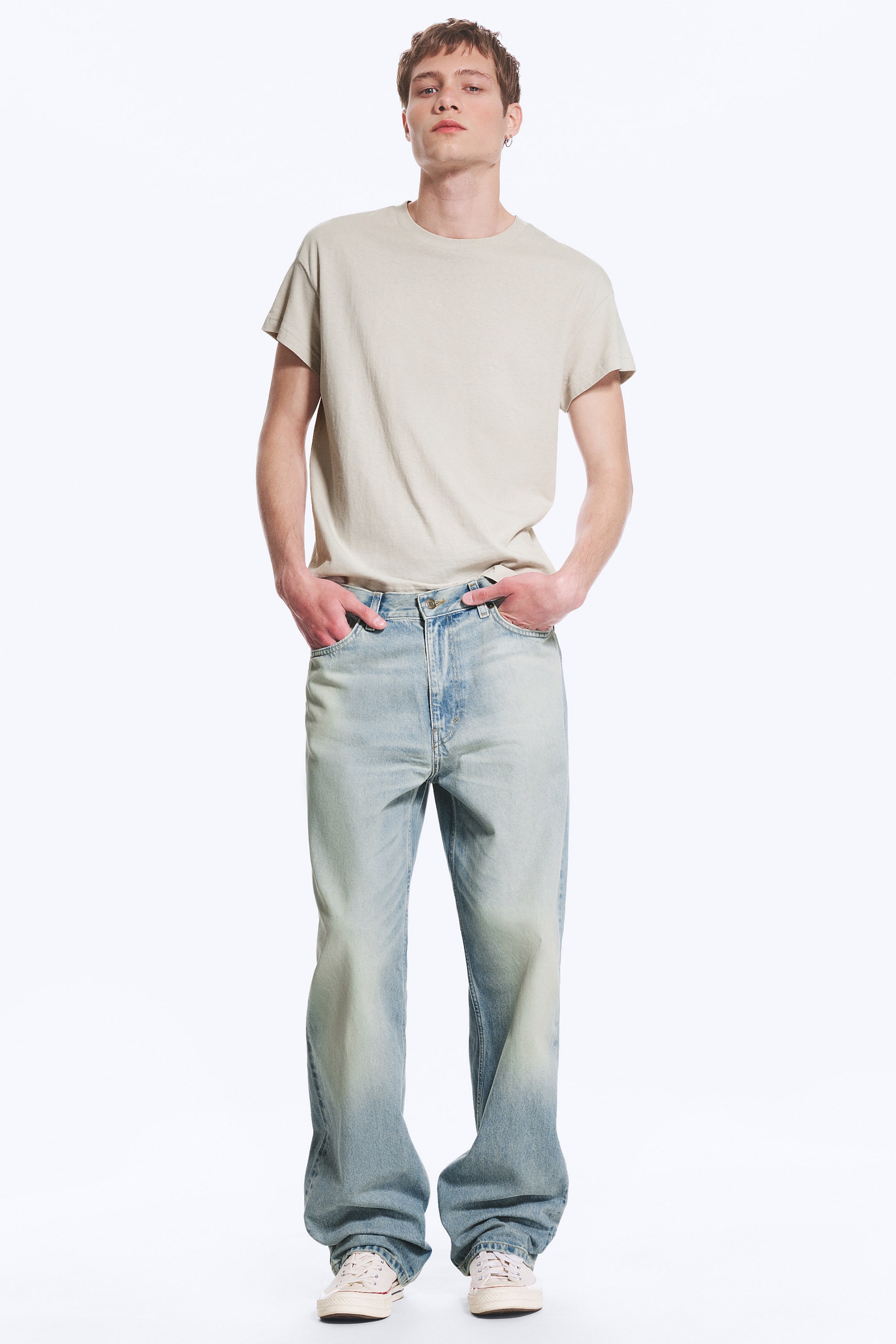 Ver imagen más grande: Una camiseta de cuello redondo en beige claro se presenta junto a unos vaqueros de pierna ancha en azul lavado claro, que presentan un degradado a un tono más oscuro en la parte inferior. Los vaqueros se amontonan sobre unas zapatillas bajas de lona ligera, con todo el conjunto mirando hacia adelante.
