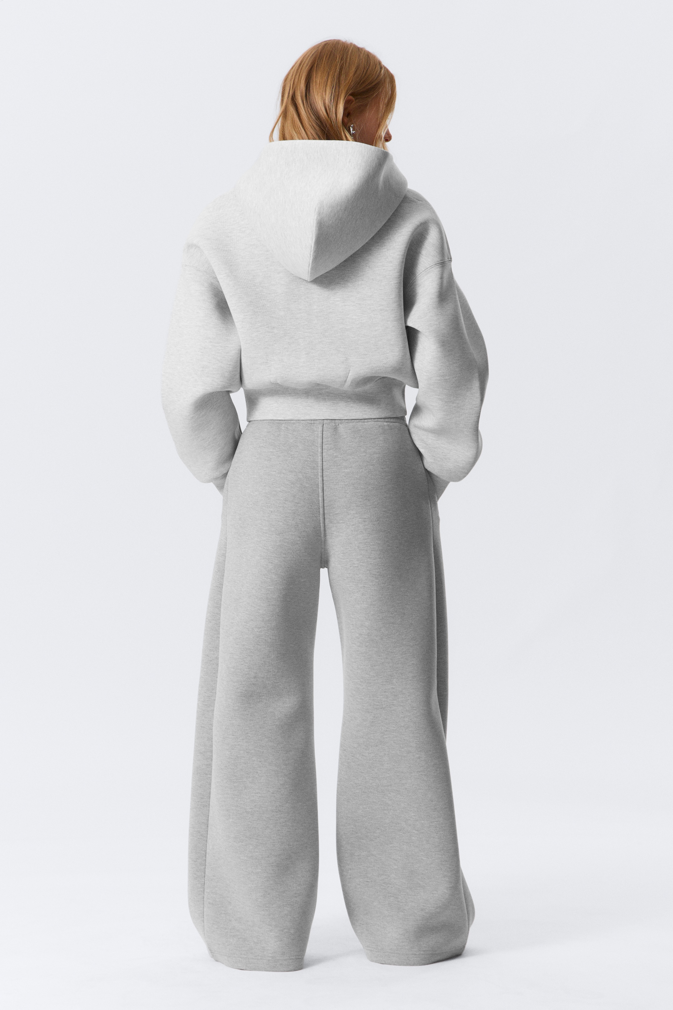 Agrandir l'image: Un sweat à capuche court en gris clair chiné et un pantalon large assorti sont présentés de dos. Le sweat à capuche présente une coupe décontractée avec une capuche, des épaules tombantes et une bande d'ourlet ajustée. Le pantalon présente une silhouette ample et fluide avec une couture centrale arrière proéminente sur chaque jambe.