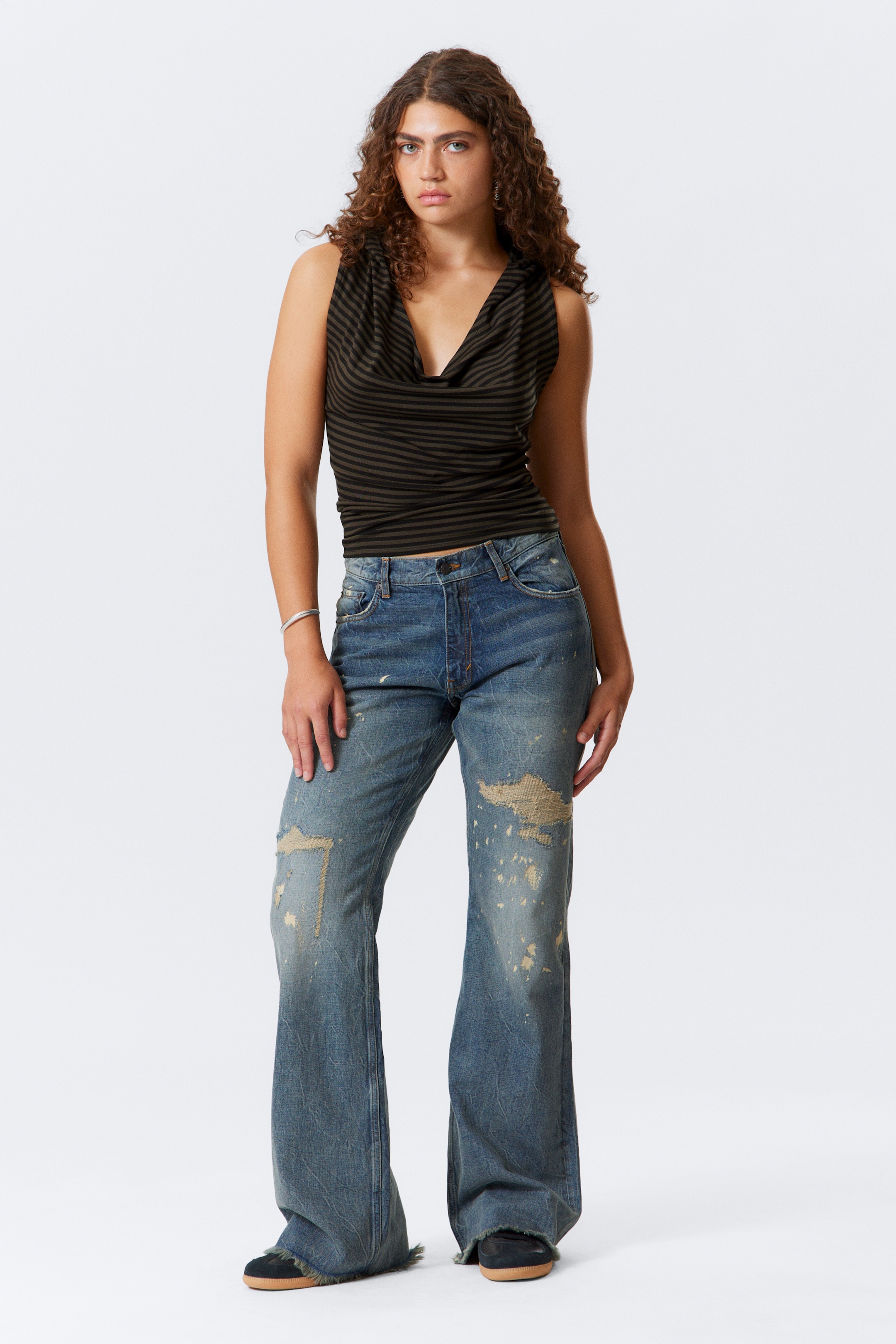 Ingrandisci l'immagine: Un top senza maniche con strisce orizzontali verde scuro e nero presenta un collo a cascata drappeggiato e una silhouette aderente. È abbinato a jeans blu lavati e strappati che mostrano toppe strappate, schizzi di vernice e una gamba ampia e svasata.