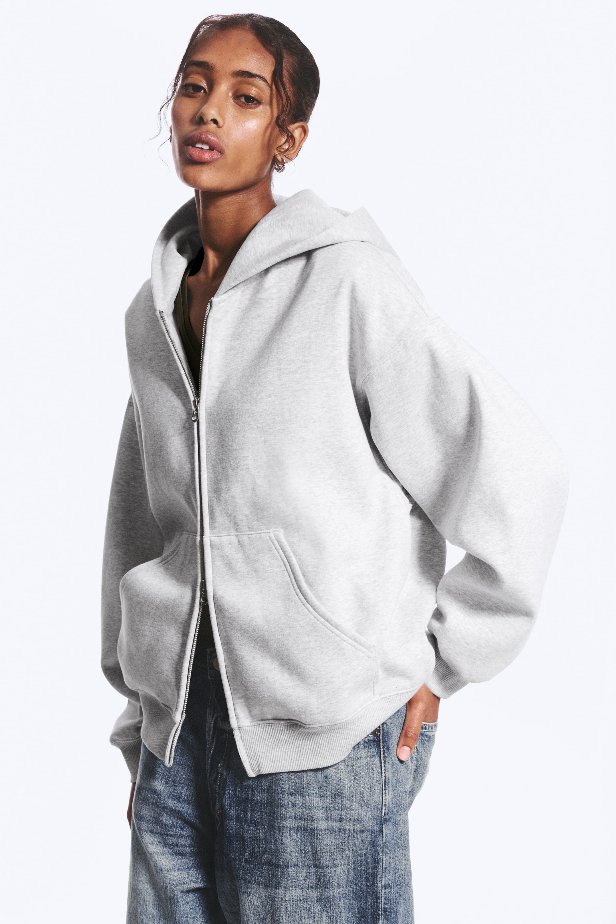 Visa större bild: En överdimensionerad, slapp ljusgrå zip-hoodie, med en rymlig dubbellager huva utan dragsnören, nedhasade axlar, en delvis uppdragen tvåvägsdragkedja, stora framfickor samt ribbade muddar och fåll, bärs av modellen. Modellen stoppar ena handen i en ficka, och en mörk topp syns under hoodien, stylad med blå jeans.