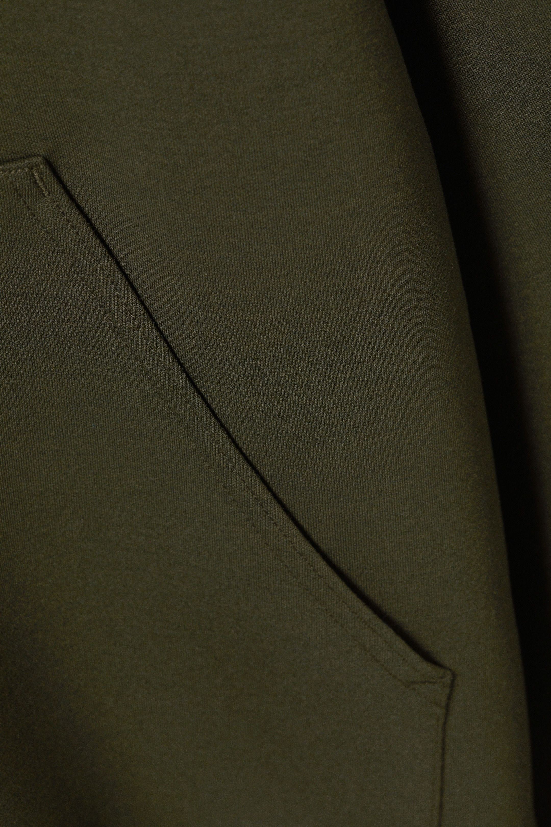 Größeres Bild anzeigen: Ein dunkelgrüner Scuba-Hoodie zeigt eine Nahaufnahme seiner Vorderseite, die eine diagonal geschnittene Kängurutasche mit sichtbaren Nähten hervorhebt.