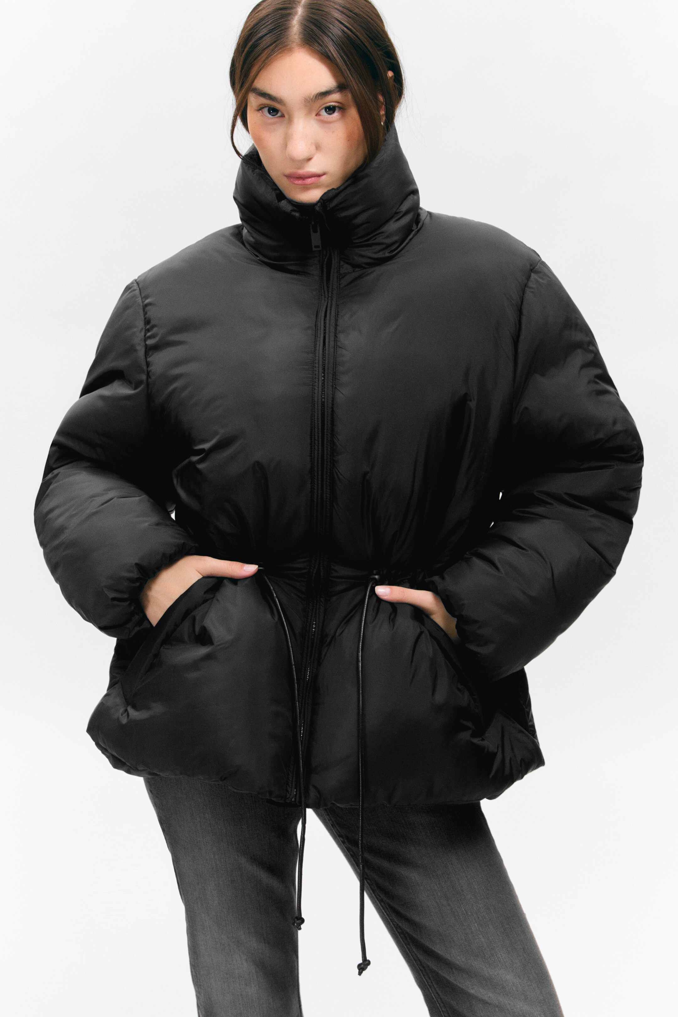 Agrandir l'image: Une veste matelassée noire est présentée de face, avec un col montant, une fermeture éclair centrale sur le devant, et une taille cintrée avec des cordons de serrage visibles. Les mains reposent dans ses poches latérales.