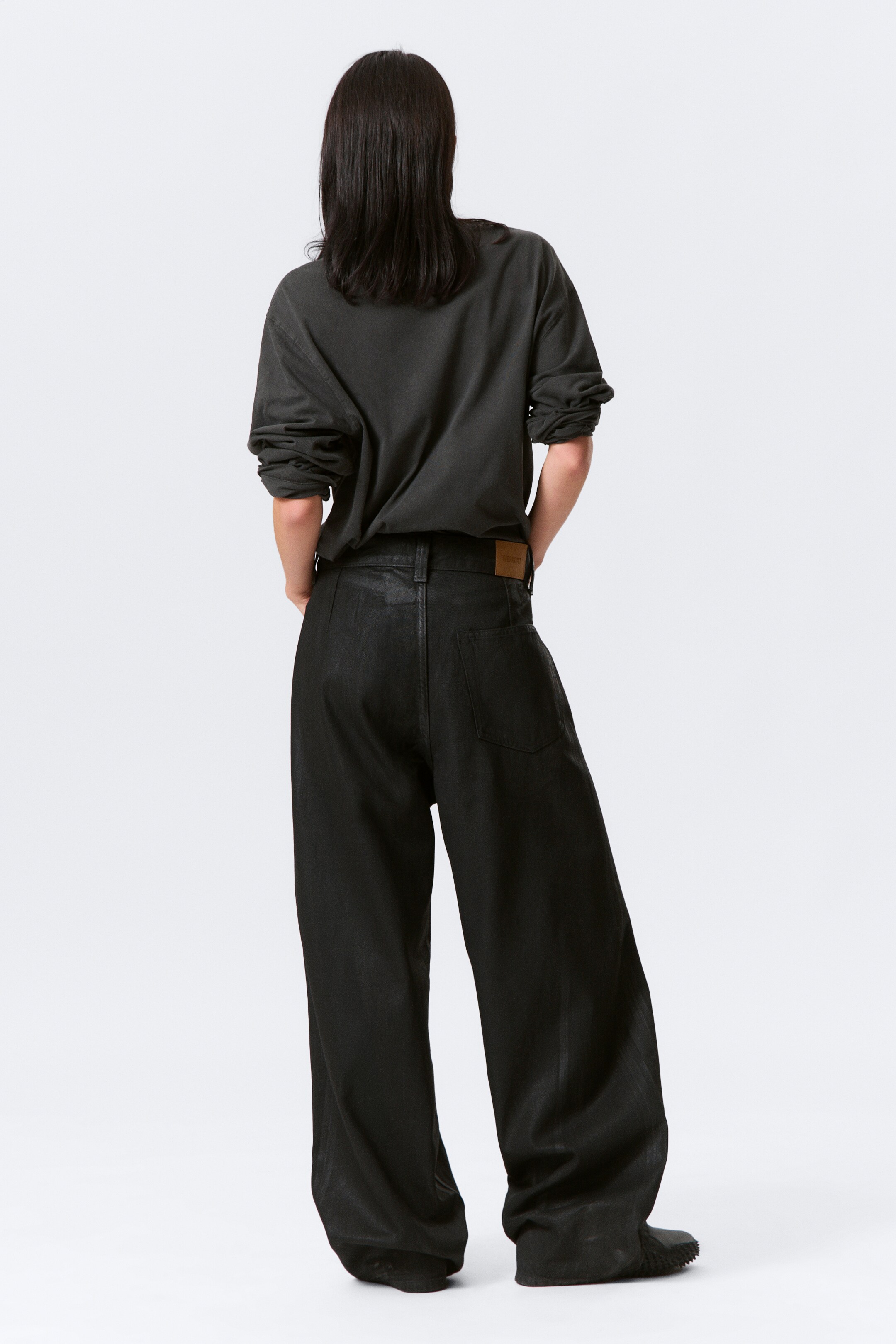 View larger image: A dark, long-sleeved t-shirt with gathered sleeves is being worn, viewed from the back, and is partially tucked into high-waisted, black wide-leg pants, which are featuring a subtle coated finish, back patch pockets, and a light brown brand patch on the waistband, with the legs pooling at the ankles.