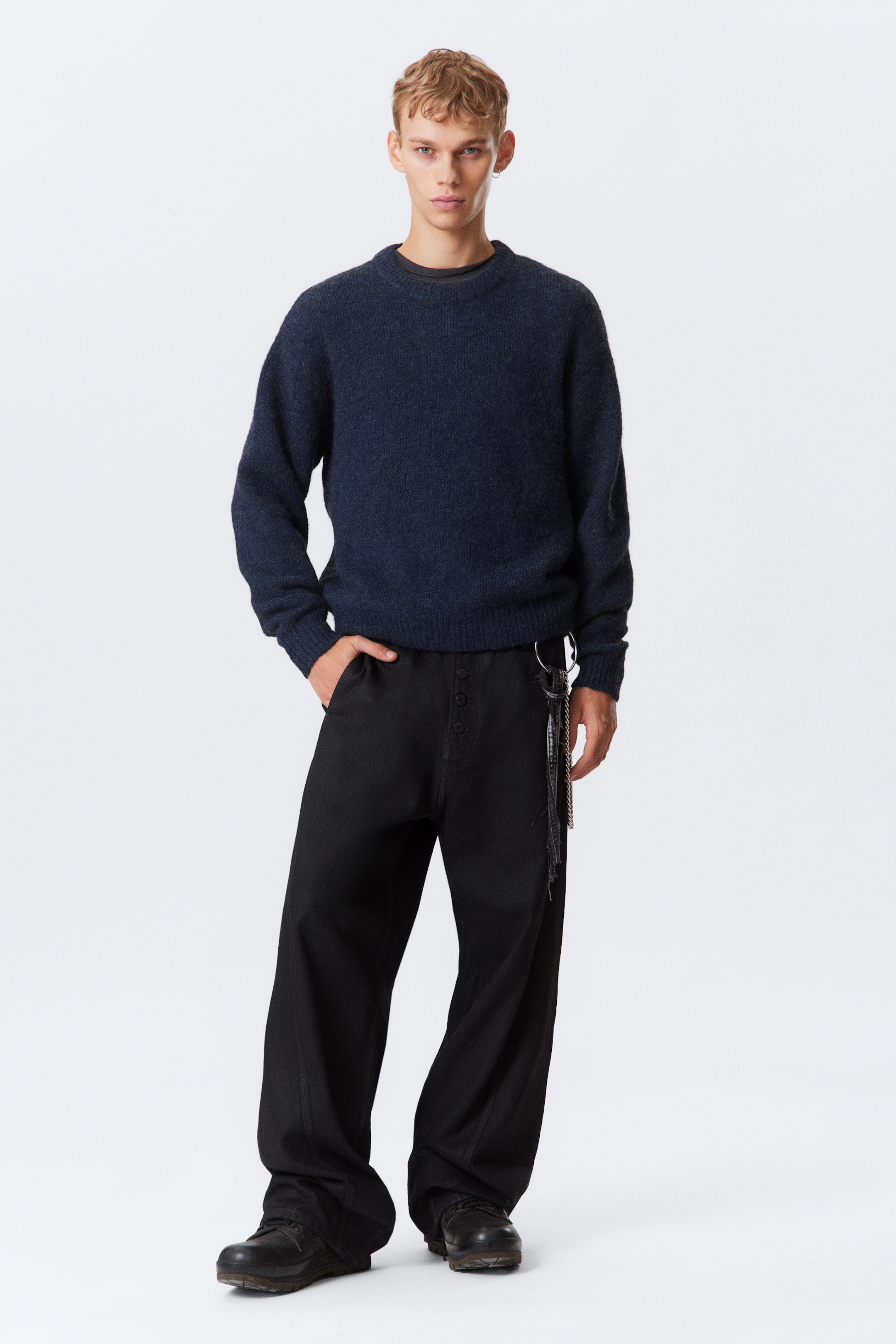 Ver imagen más grande: Un suéter de cuello redondo azul oscuro, con una textura suave y un ajuste relajado, se está usando sobre una camiseta oscura. También se están usando unos pantalones negros de pierna ancha, que muestran una bragueta de botones visible y un accesorio de cadena de metal que cuelga de la cadera derecha.