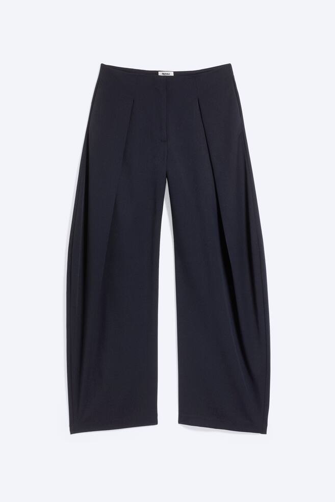 Low-rise balloon trousers in dark navy blue are shown from a front-facing angle, featuring a wide, curved-leg silhouette with deep front pleats that create a voluminous shape. The trousers feature a flat waistband and straight, wide hems. - 1