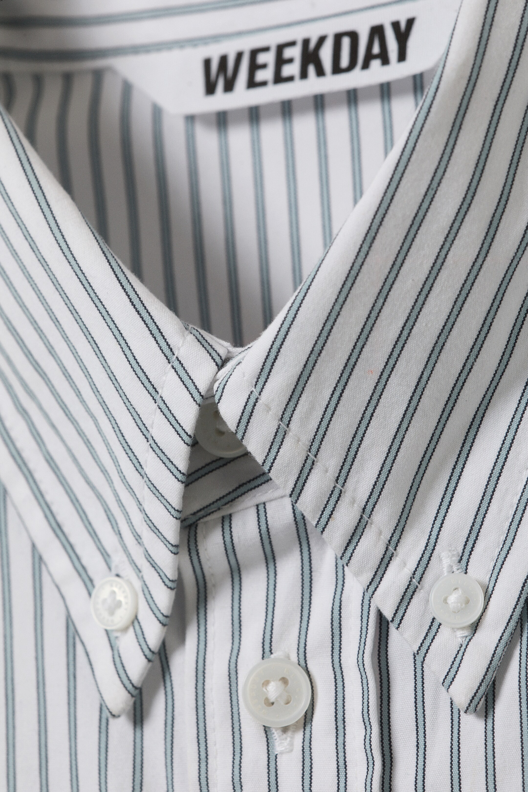 Ver imagem maior: Uma camisa branca com colarinho, mostrada de frente, é padronizada com finas riscas verticais pretas e azul claro. Os pontos do colarinho com botões estão presos com botões brancos, e botões brancos correspondentes estão a segurar a carcela central, com a etiqueta da marca 'WEEKDAY' visível no colarinho interno.