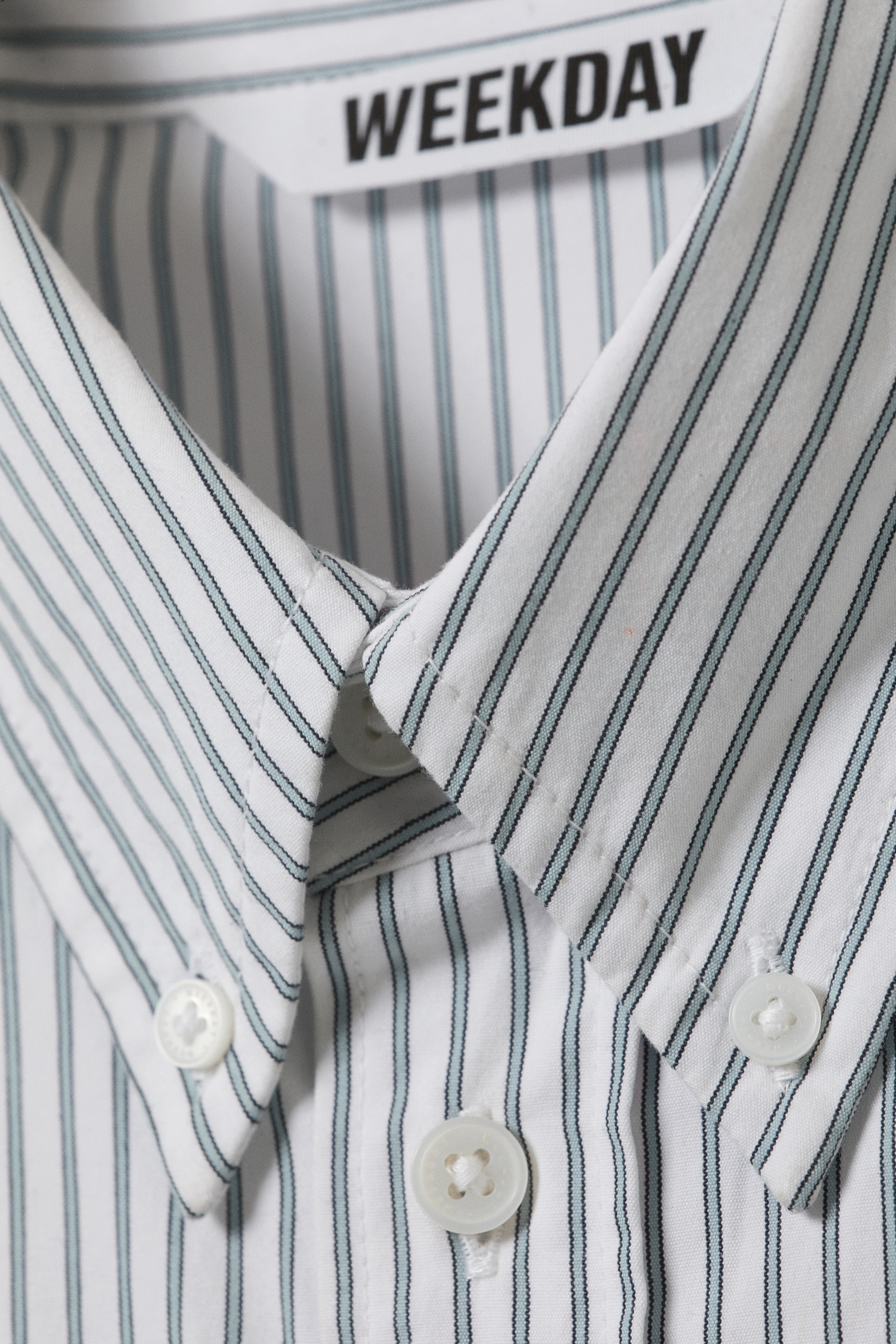 View larger image: A white collared shirt, shown from the front, is patterned with thin black and light blue vertical stripes. Its button-down collar points are fastened with white buttons, and matching white buttons are securing the central placket, with the 'WEEKDAY' brand tag visible on the inner collar.