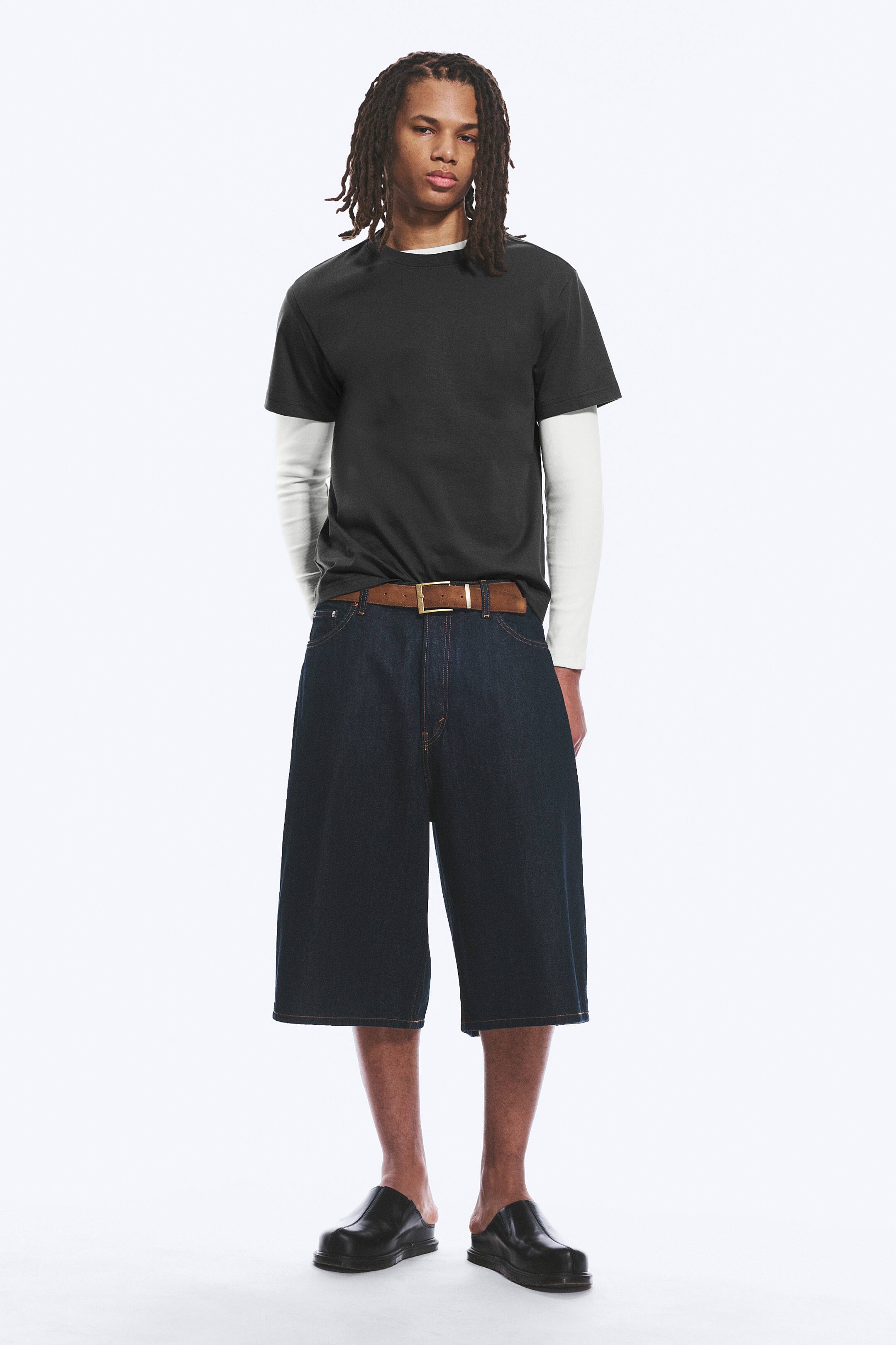 View larger image: A dark grey short-sleeved t-shirt is layered over a white long-sleeved shirt. Wide-leg dark wash denim shorts are worn, secured by a brown belt with a gold buckle, and black slip-on mules complete the ensemble.