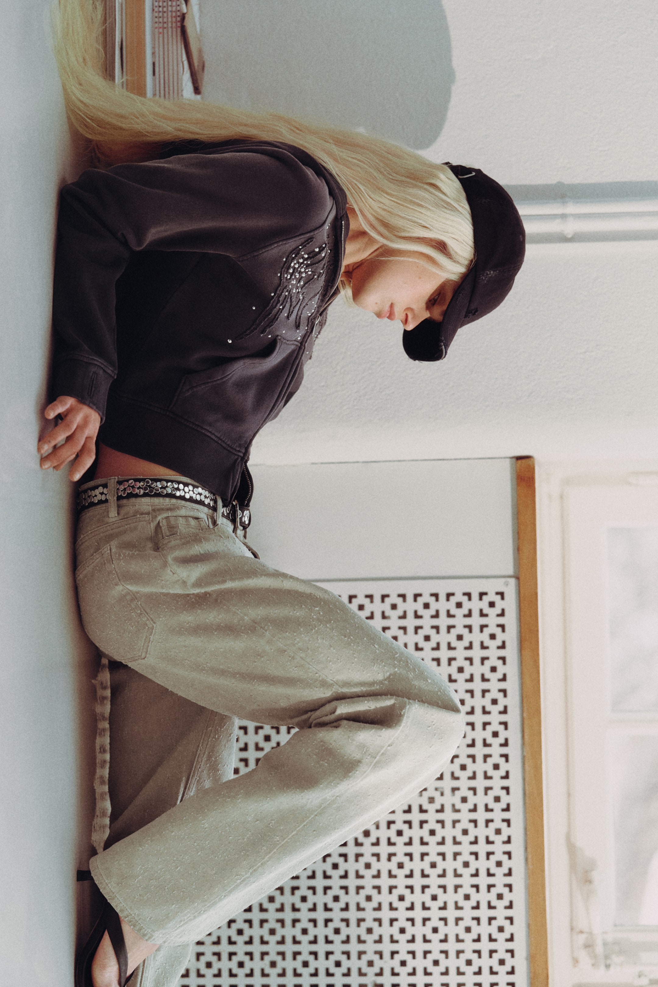 Ingrandisci l'immagine: Una felpa con cappuccio grigio scuro con chiusura a zip è presentata, caratterizzata da una chiusura frontale con cerniera e maniche lunghe con polsini e orlo a coste. L'area del petto sinistro anteriore presenta un brillante design a forma di ala con borchie.