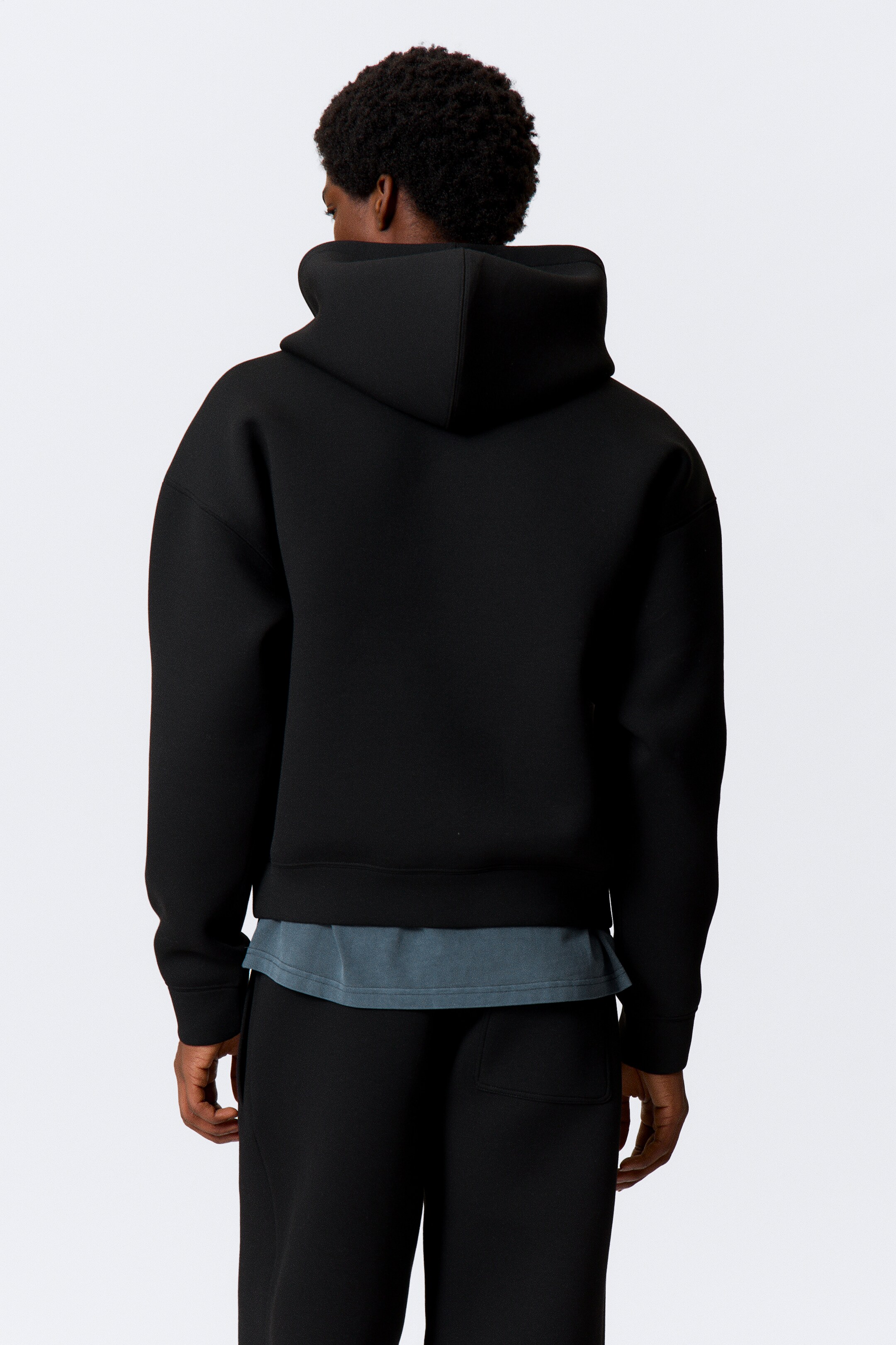 Grösseres Bild ansehen: Ein schwarzer Kapuzenpullover wird von hinten gezeigt, mit einer strukturierten Kapuze und locker geschnittenen Ärmeln. Er besteht aus einem glatten, strukturierten Stoff, mit gerippten Bündchen und Saum, und ein hellblaues T-Shirt wird unter seinem Saum getragen.