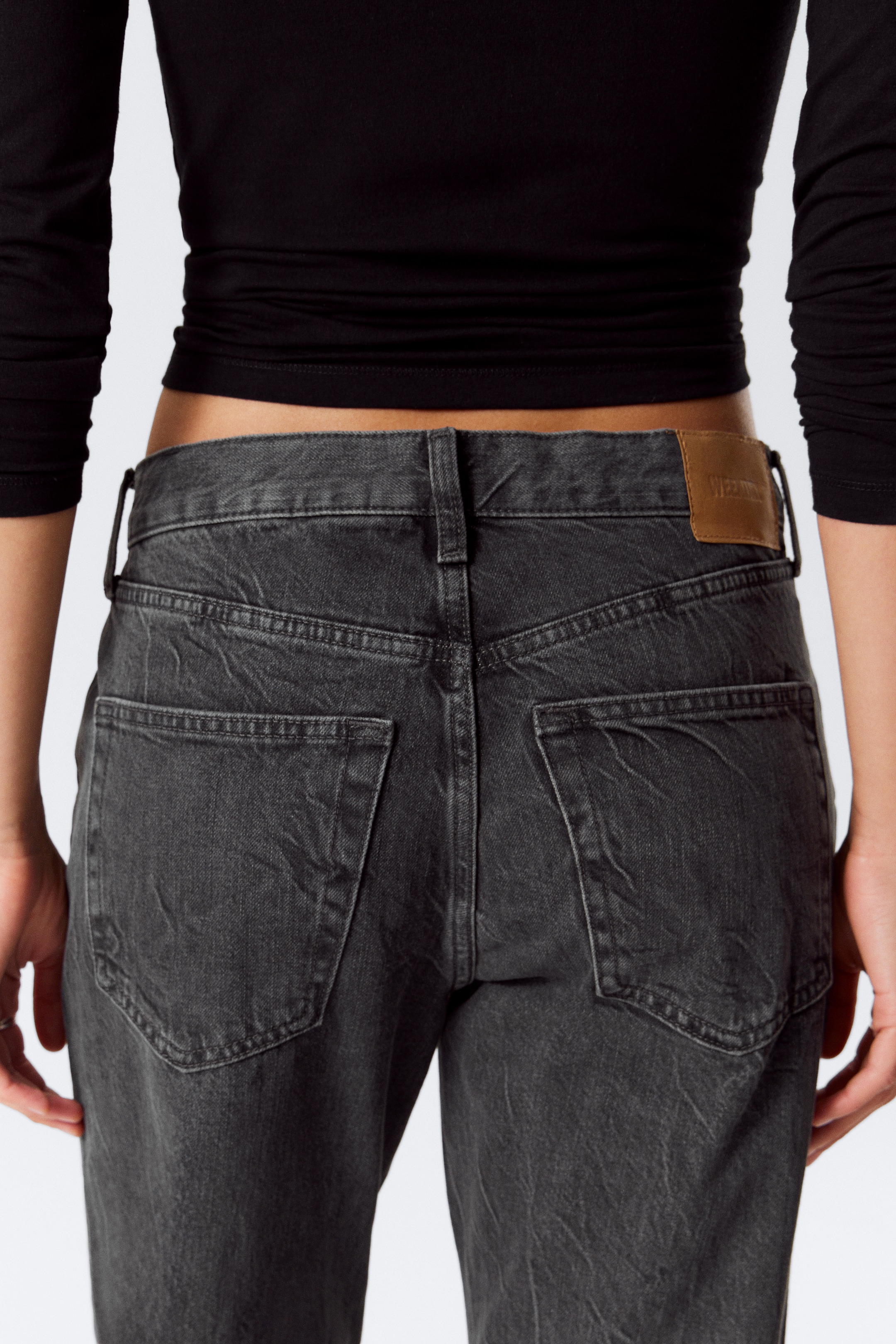 Grösseres Bild ansehen: Dunkelgraue Denim-Jeans werden von hinten betrachtet, mit zwei dreieckigen Gesäßtaschen und einem hellbraunen Markenaufnäher auf der rechten Seite des Bunds. Die Jeans weisen eine verblasste, zerknitterte Textur auf und werden mit einem schwarzen langärmeligen Oberteil getragen, das knapp über der Taille sichtbar ist.