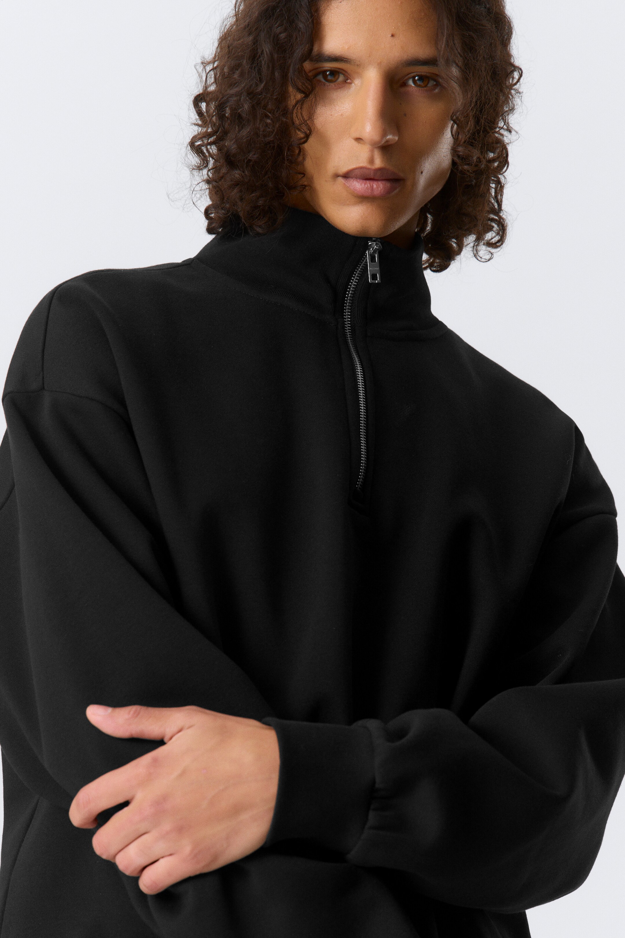 Ver imagen más grande: Un jersey negro con media cremallera presenta un cuello alto con una cremallera parcialmente abierta de tono plateado. Presenta un ajuste relajado con mangas largas y puños acanalados, mostrado desde una vista frontal ligeramente angular.