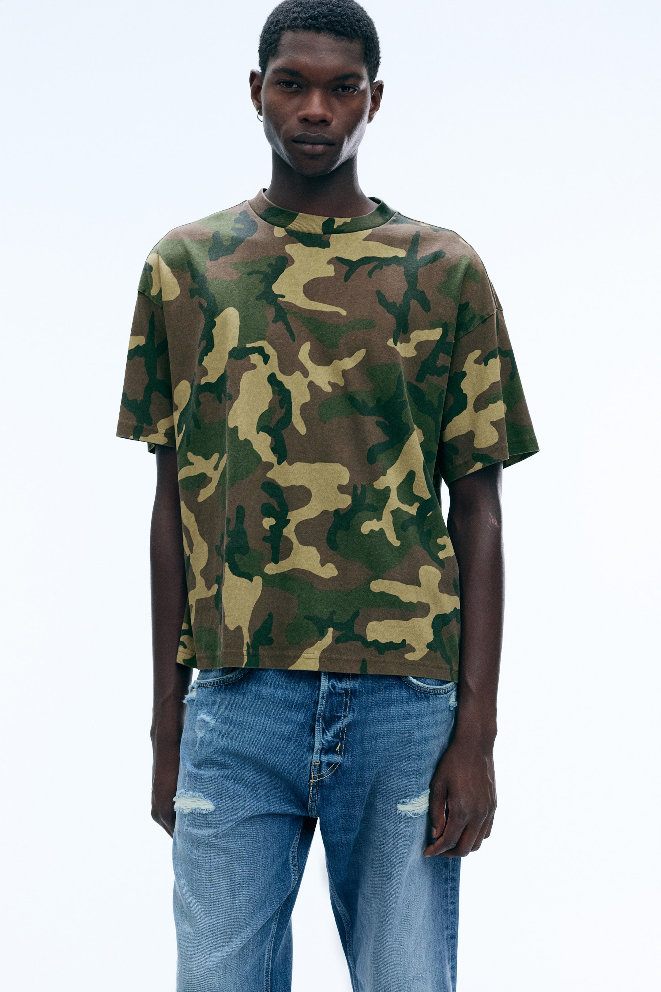 Größeres Bild ansehen: Ein locker sitzendes T-Shirt mit Camouflage-Print, kastiger Silhouette, Rundhalsausschnitt und überschnittenen Schultern wird vom Model getragen. Das kurzärmelige Oberteil ist mit Jeans in Distressed-Optik kombiniert.