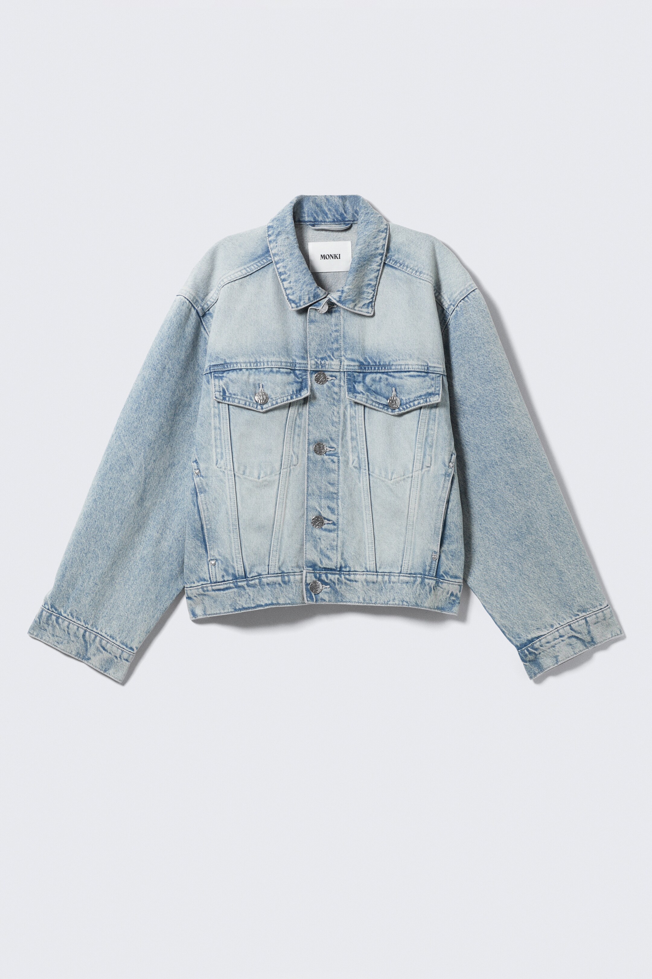 View larger image: A light blue faded denim jacket is presenting its front view, featuring a pointed collar with a "MONKI" brand label. It is showcasing a full button-front closure with metal buttons, two chest flap pockets with button closures, and two side welt pockets. The jacket is designed with long sleeves ending in buttoned cuffs and is displaying an oversized fit.