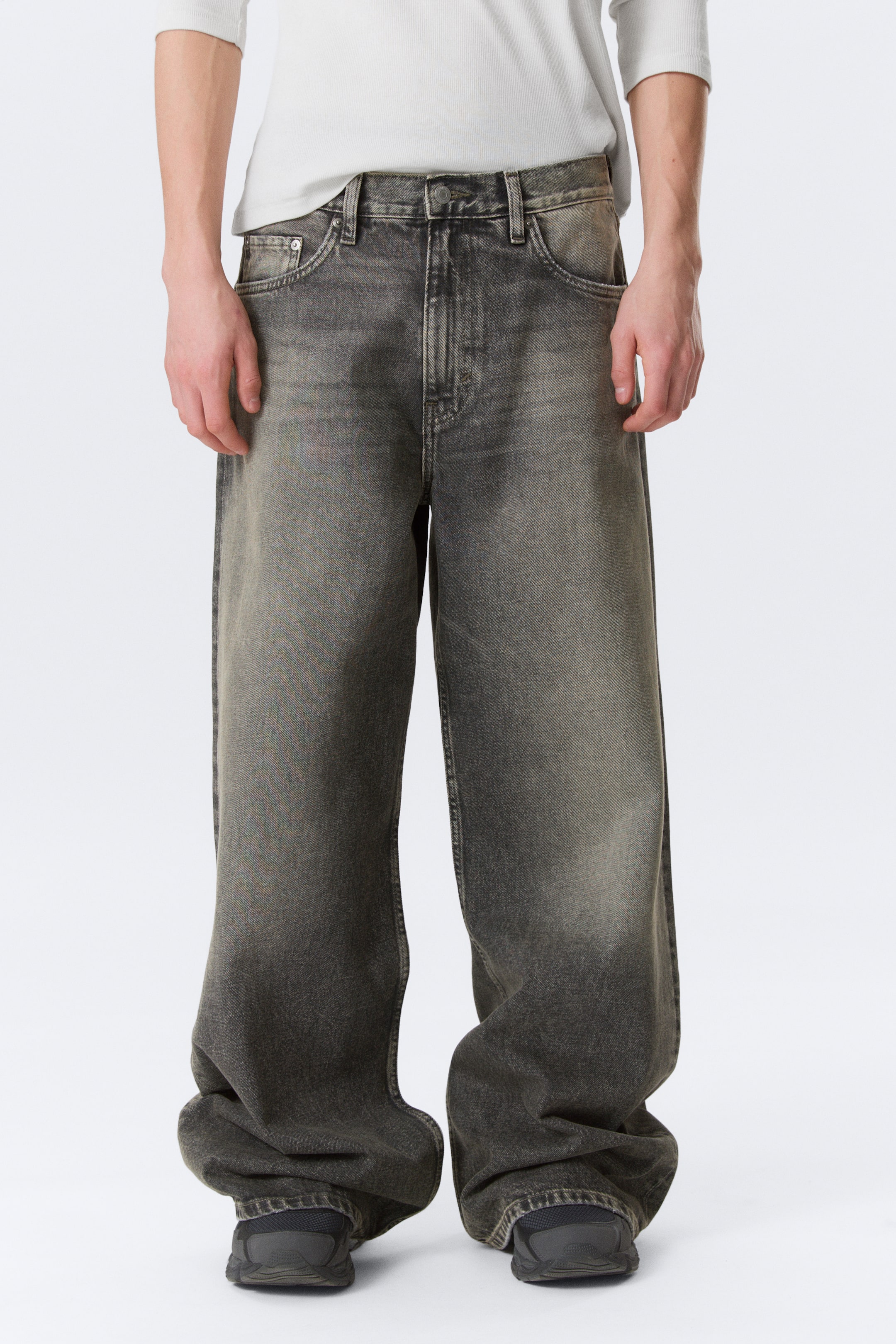 Ver imagem maior: Jeans cinza escuro de perna larga apresentam um efeito desbotado e um corte solto, vistos de uma perspetiva frontal. Exibem um fecho de botão e fecho éclair, presilhas para cinto, bolsos frontais em concha e estão a acumular-se na bainha sobre sapatos escuros.