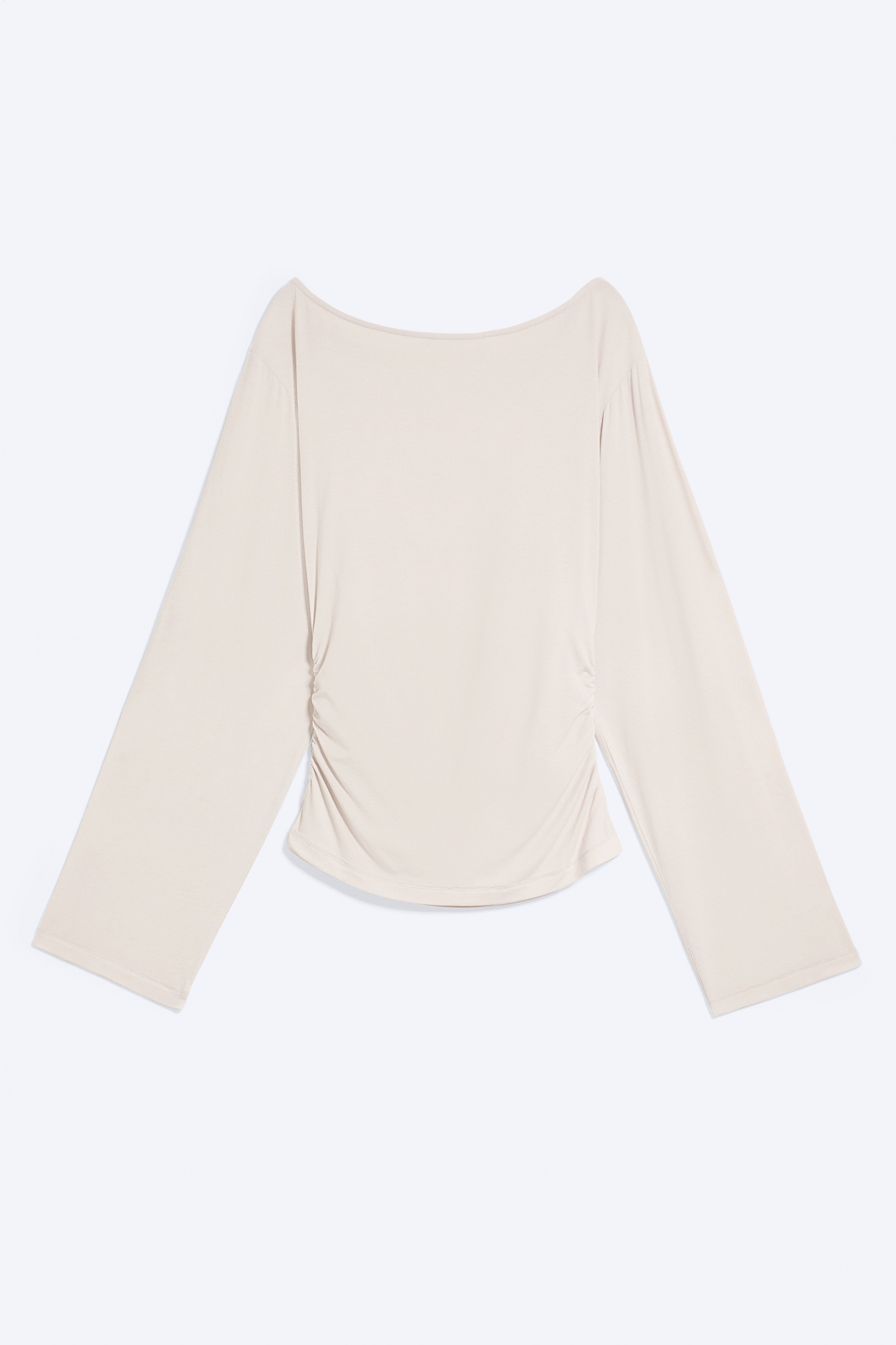Un haut beige clair à manches longues, doté d’un col bateau, de manches amples et d’une taille ajustée froncée, est porté par le mannequin. Le mannequin associe ce haut à un pantalon texturé gris foncé, les mains glissées dans les poches.