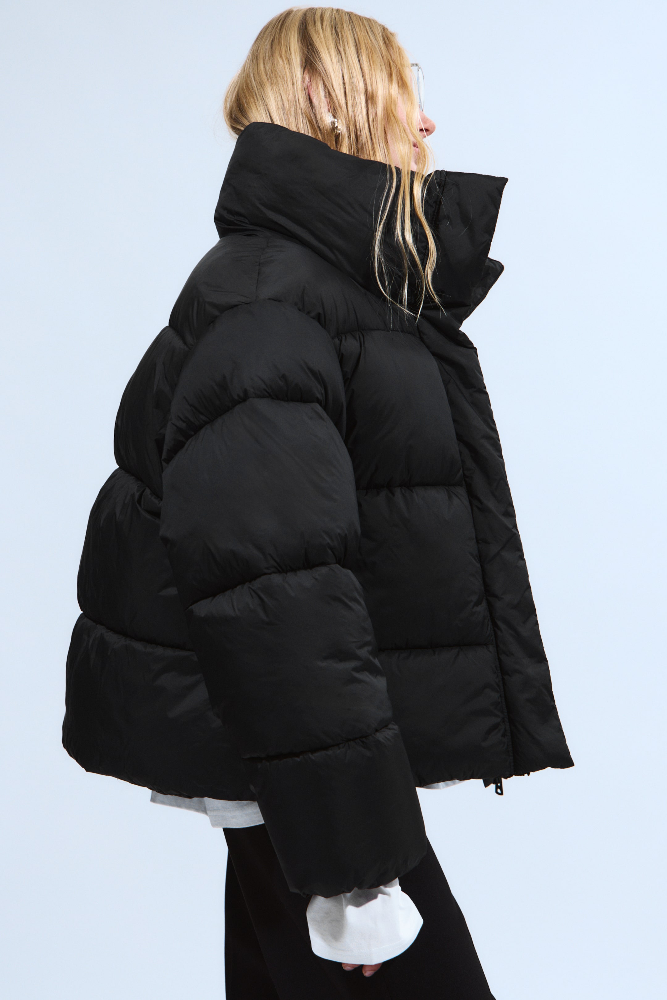 Ingrandisci l'immagine: Una giacca a vento nera è presentata di profilo laterale, rivolta a destra, caratterizzata da una vestibilità oversize con prominenti cuciture orizzontali e un alto colletto rialzato. Un polsino di camicia bianca è visibile che si estende da sotto la manica della giacca.