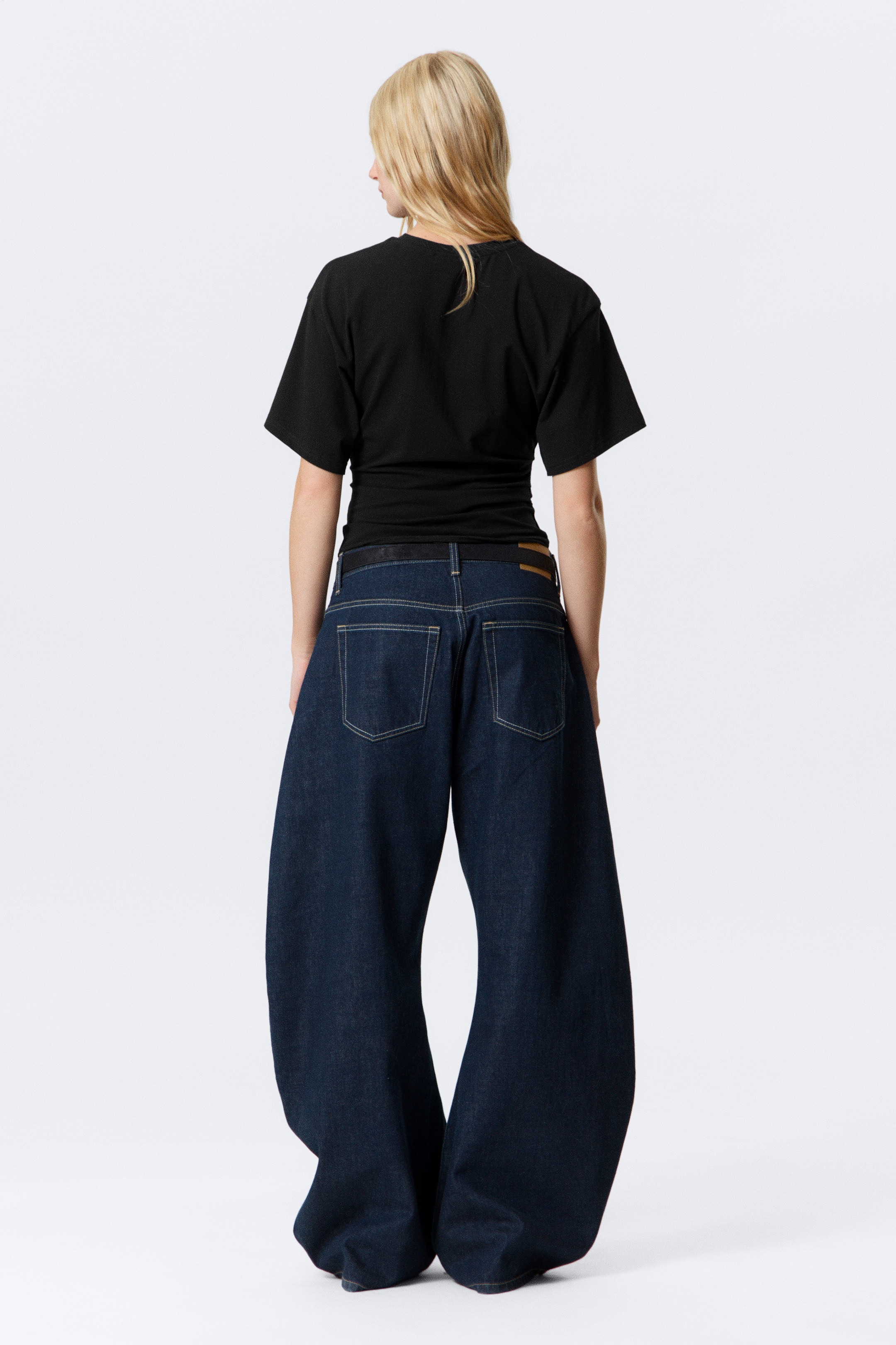 Grösseres Bild ansehen: Ein schwarzes kurzärmeliges Oberteil und dunkel gewaschene Jeans mit weitem Bein werden von hinten gezeigt. Das Oberteil ist an der Taille gerafft, während die Jeans zwei aufgesetzte Gesäßtaschen, helle Kontrastnähte und ein extrem weites Bein aufweisen, das sich an den Knöcheln staut. Ein dunkler Gürtel und ein hellbrauner Markenaufnäher sind ebenfalls am Bund der Jeans zu sehen.