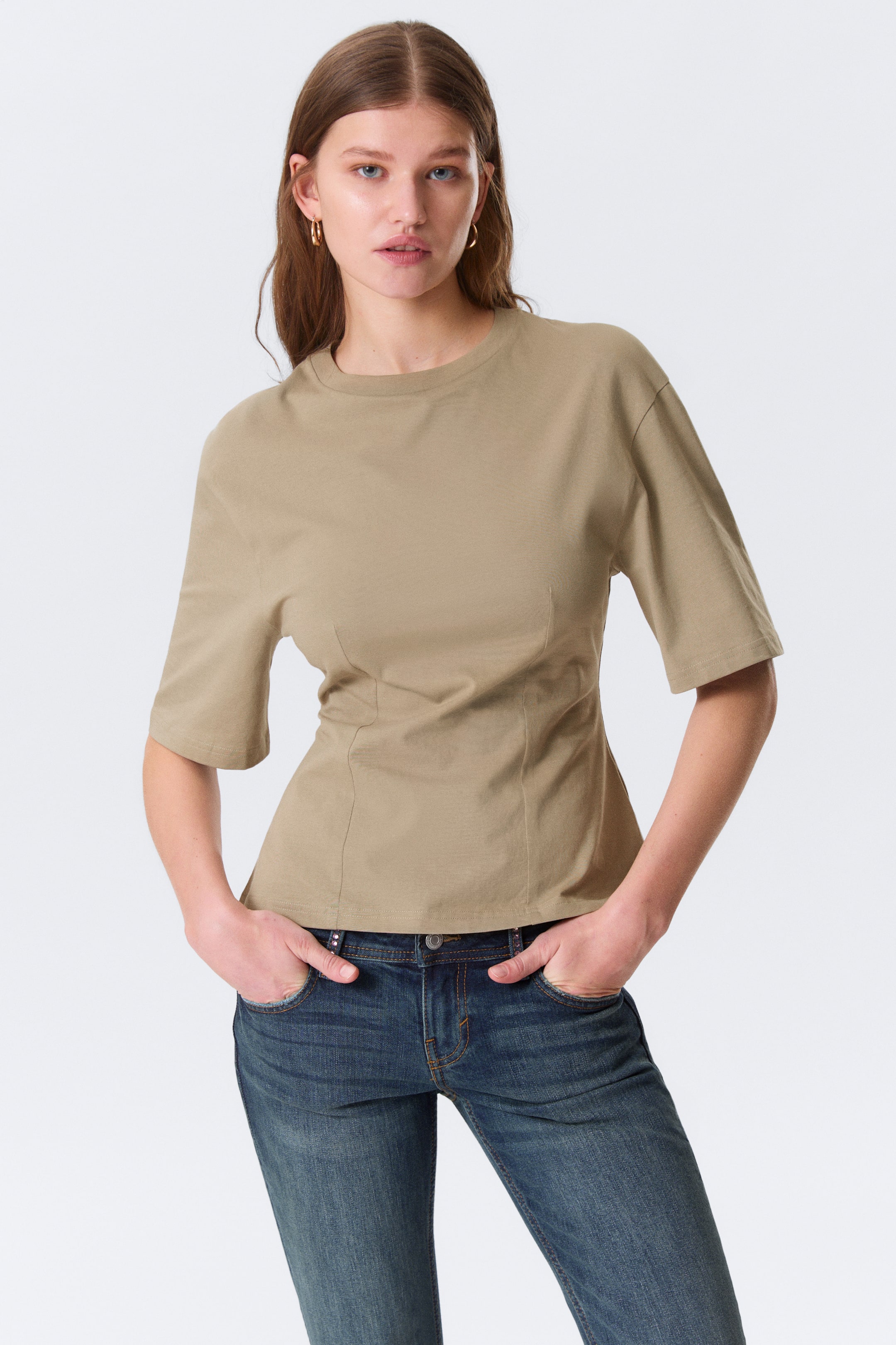 Visualizza immagine più grande: Un top beige presenta un girocollo e maniche rilassate lunghe fino al gomito. È progettato con cuciture verticali in vita, creando una silhouette aderente che si allarga leggermente all'orlo, cadendo sciolto sopra i jeans blu.